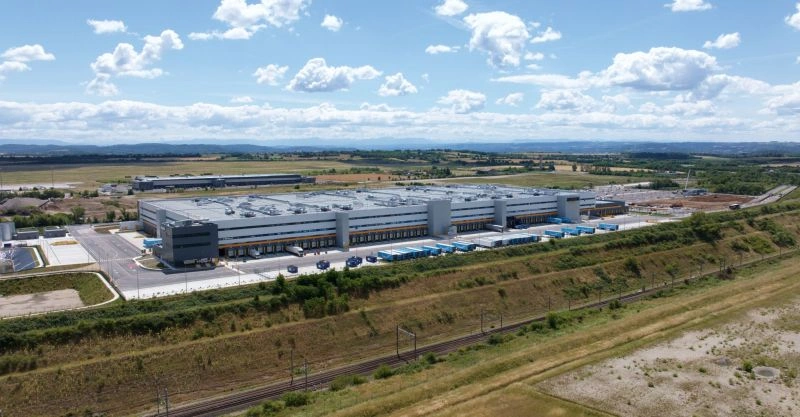 Vue aérienne d'un grand entrepôt logistique avec des camions bleus aux quais sous un ciel nuageux.