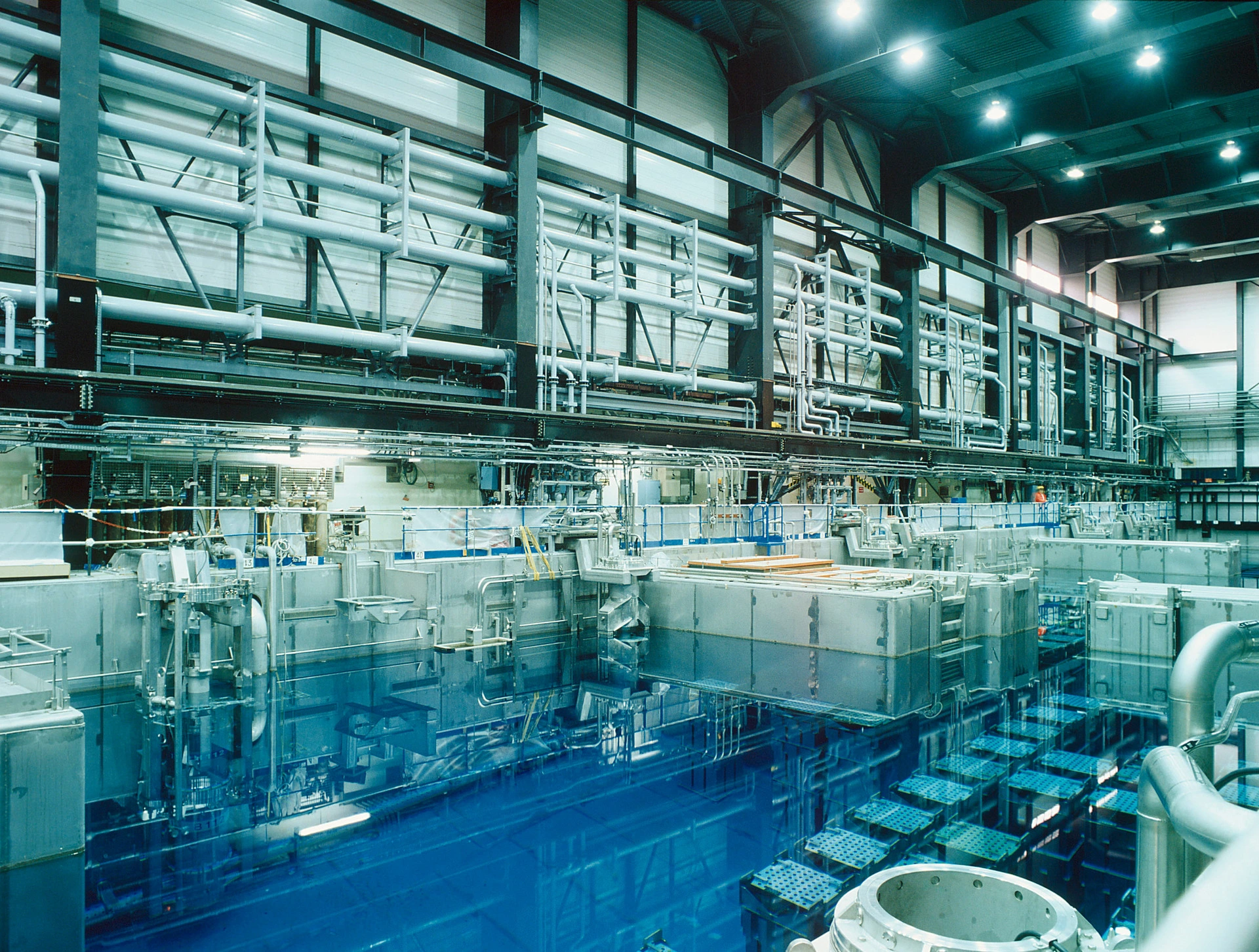 Intérieur d'une installation nucléaire montrant une grande piscine d'eau bleu vif avec des structures submergées, entourée de machines métalliques grises et de tuyauteries aériennes complexes.