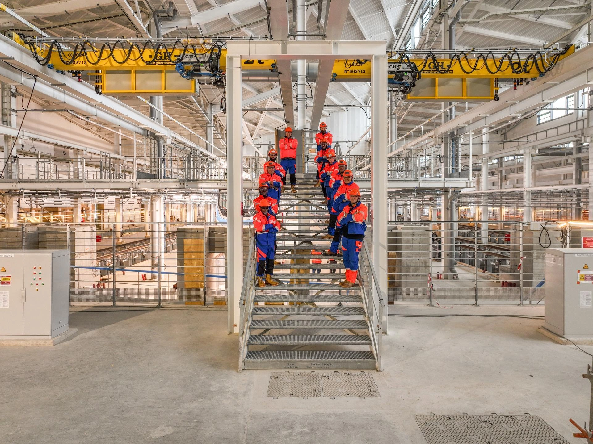 Dix ouvriers souriants en uniformes de travail orange et bleus, coiffés de casques de sécurité orange, posent sur un escalier métallique au centre d'un vaste hall industriel moderne et lumineux, équipé de ponts roulants jaunes.