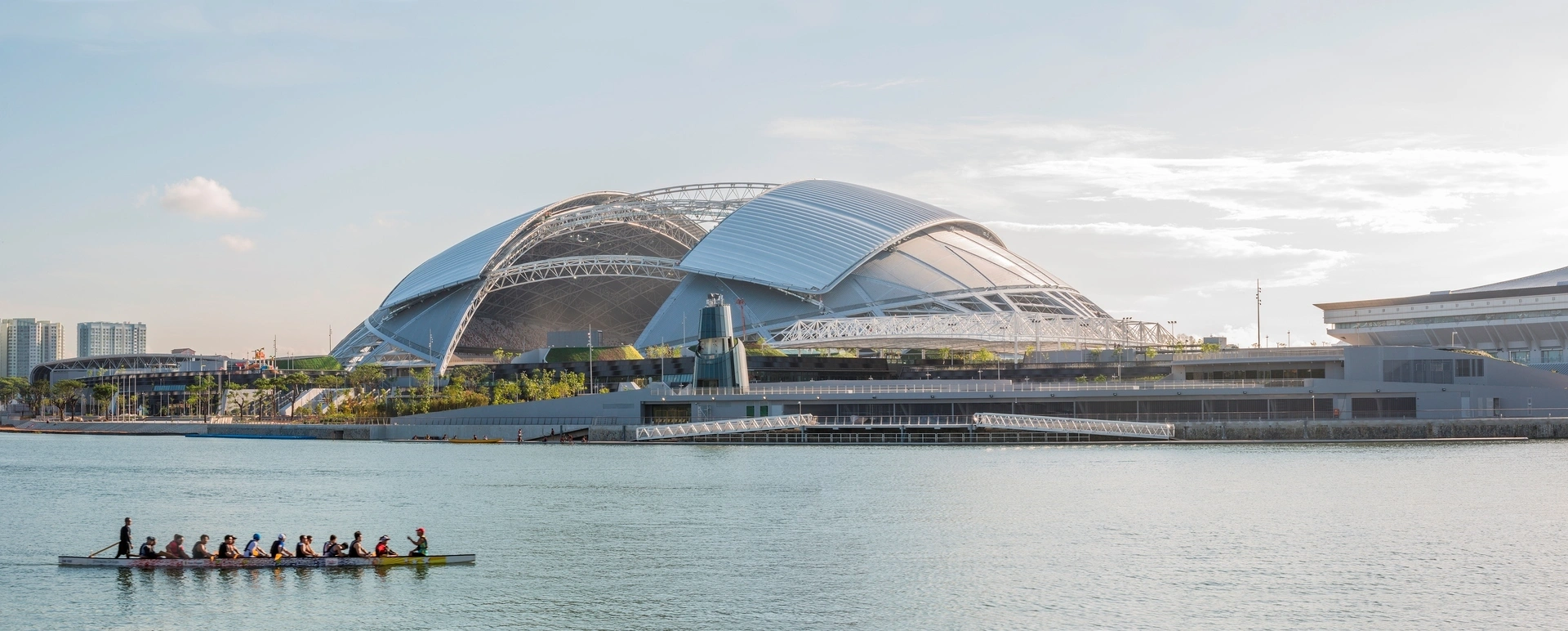 Des dragon boaters rament sur des eaux calmes et bleues devant le moderne Singapore Sports Hub au dôme argenté et ses aménagements paysagers verts, sous un ciel clair.