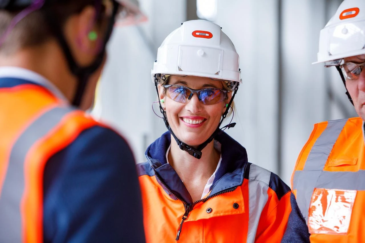 Une femme souriante porte un casque blanc, des lunettes de sécurité réfléchissantes et une veste de travail orange et bleue avec des bandes réfléchissantes, regardant un collègue. Elle a des bouchons d'oreille verts et un micro-casque, avec deux autres travailleurs en tenue de sécurité similaire partiellement visibles autour d'elle.