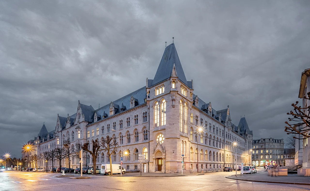 Un grand bâtiment historique en pierre claire de style néo-gothique, avec des toits en ardoise grise et des fenêtres éclairées, domine une rue urbaine au crépuscule. Le ciel est couvert de nuages sombres, et les lampadaires projettent une lumière chaude sur l'asphalte.