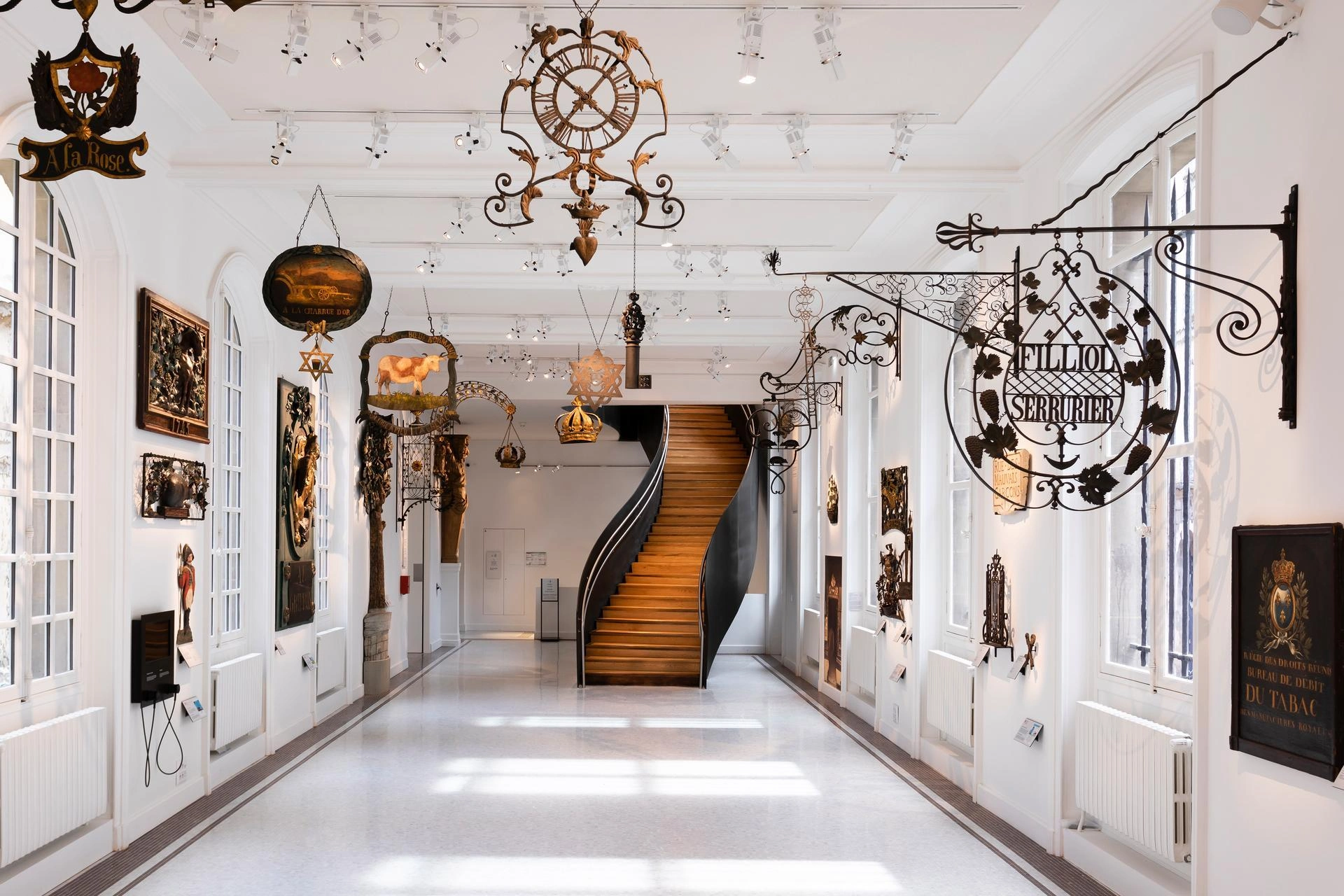 Une lumineuse galerie de musée présente une collection d'enseignes de magasins anciennes en fer forgé et en bois sculpté, suspendues au plafond et fixées aux murs blancs. Un élégant escalier en colimaçon sombre avec des marches en bois mène au fond du long couloir.