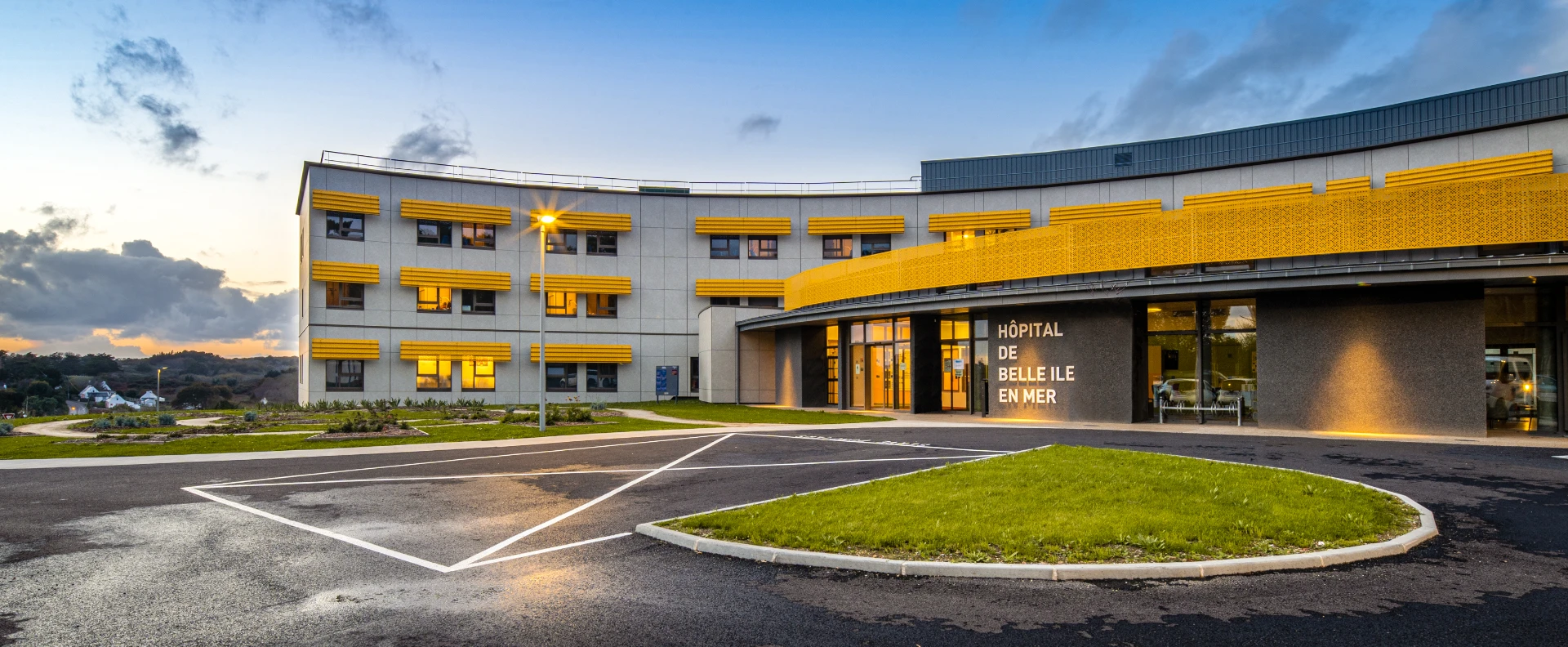 Le bâtiment incurvé et moderne de l'Hôpital de Belle Île en Mer, avec sa façade grise et ses éléments décoratifs jaune vif, est illuminé au crépuscule. On y voit l'entrée principale, un parking et des pelouses.