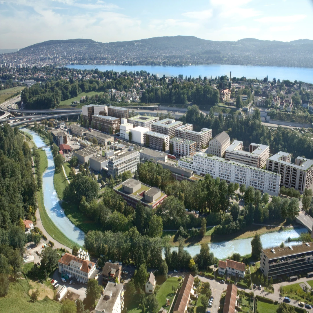Vue aérienne d'un grand projet de développement urbain moderne, avec des bâtiments aux façades variées longeant une rivière sinueuse et des parcs arborés. À l'arrière-plan, un vaste lac bleu et des montagnes verdoyantes complètent le paysage sous un ciel clair et ensoleillé.