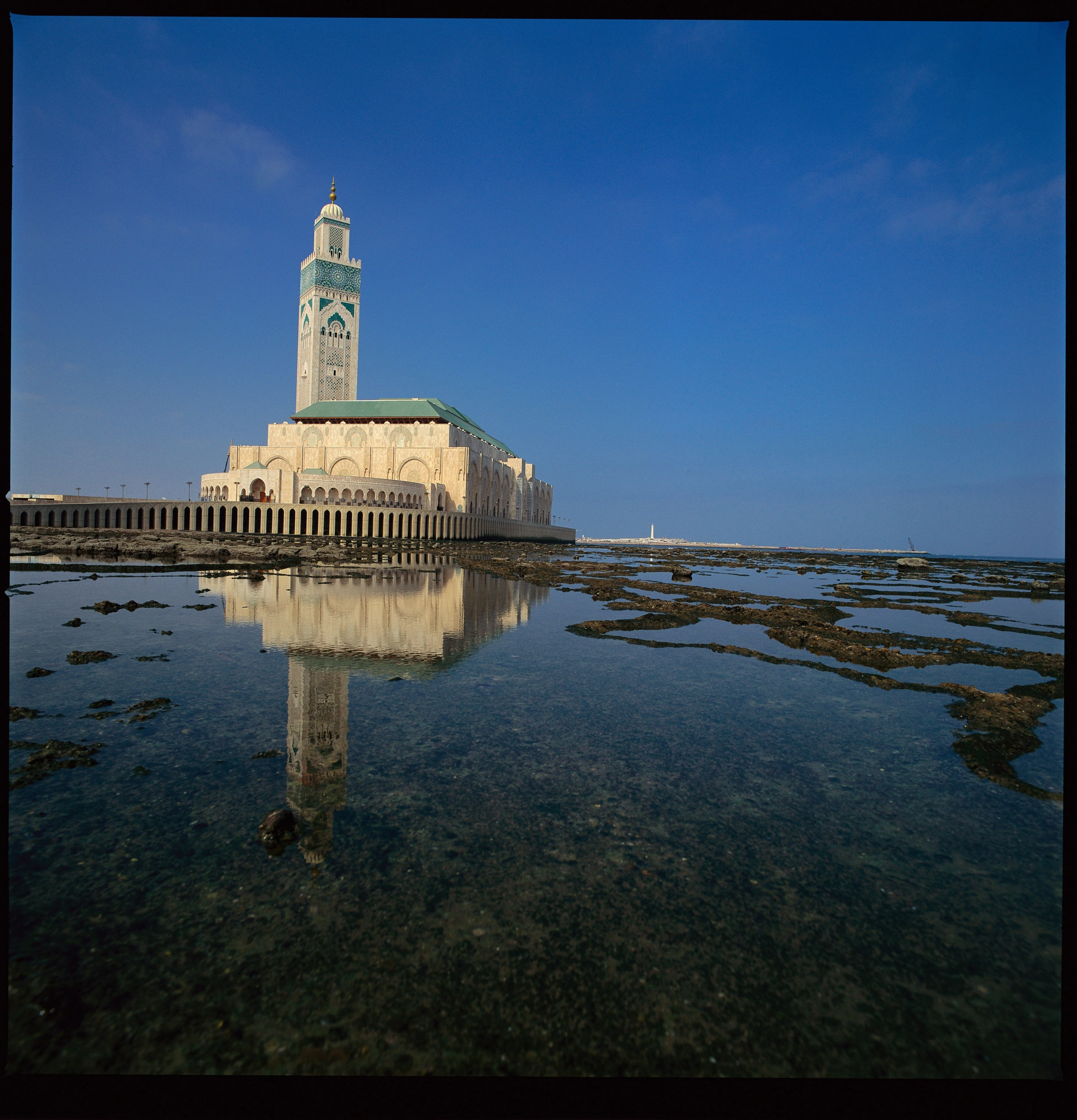 La majestueuse mosquée Hassan II de Casablanca, avec son minaret blanc et vert orné, se reflète dans l'eau claire de la côte rocheuse sous un ciel bleu intense.