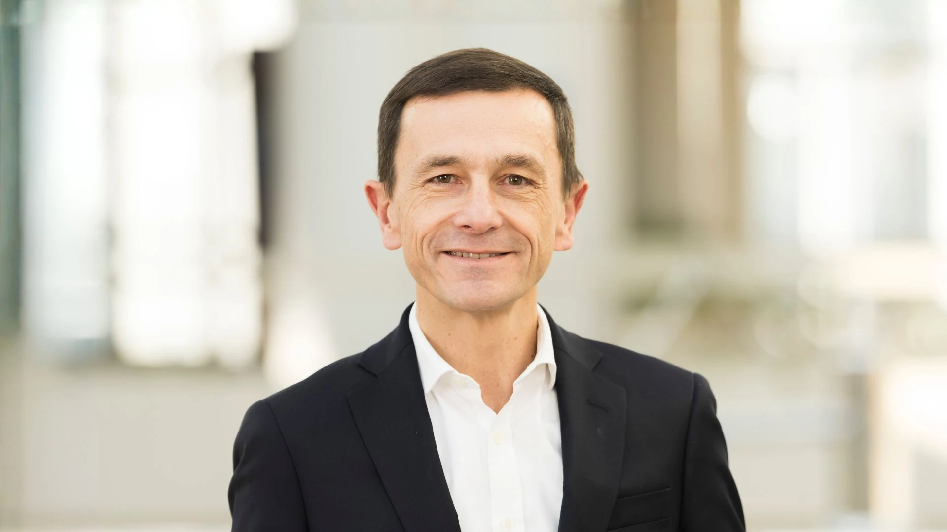 Homme d'affaires souriant d'âge moyen aux cheveux bruns et courts, vêtu d'un costume sombre et d'une chemise blanche, sur un fond lumineux et flou.
