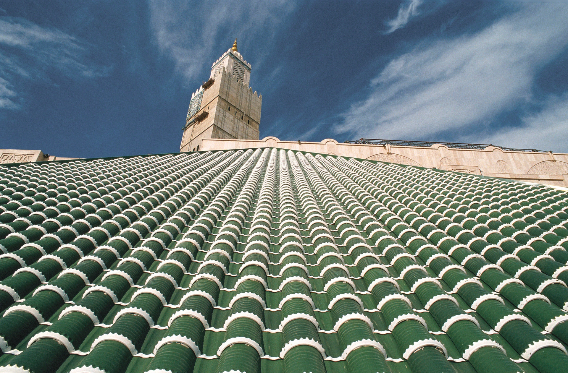 Vue en contre-plongée d'un toit orné de tuiles vertes et blanches en rangées régulières, avec un minaret de couleur claire coiffé d'or se dressant sous un ciel bleu nuageux.