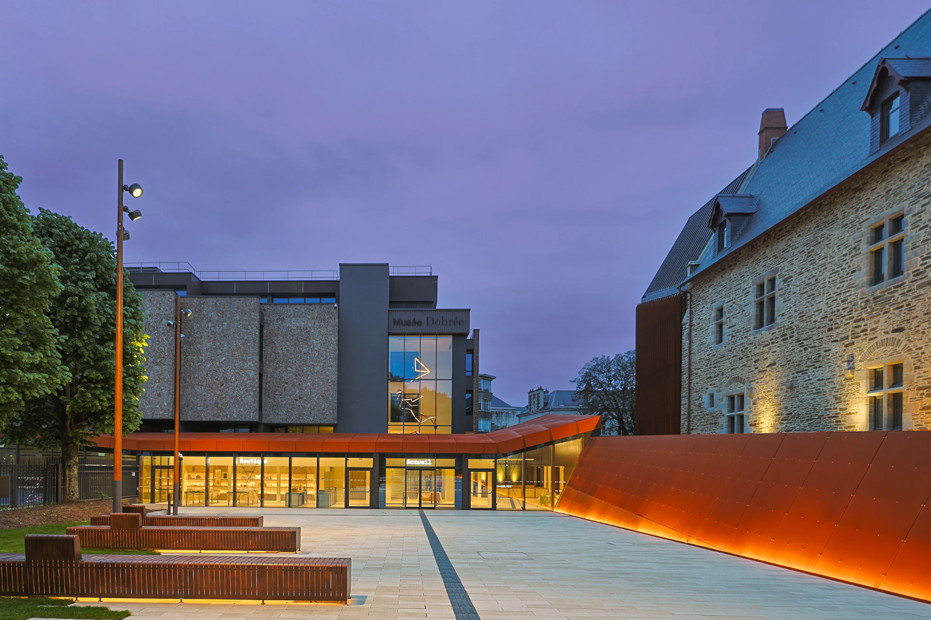 Vue extérieure du Musée Dobrée au crépuscule, combinant un bâtiment historique en pierre et une extension moderne gris et verre, avec un parvis lumineux doté de bancs et de murs orange-rouille sous un ciel teinté de violet.