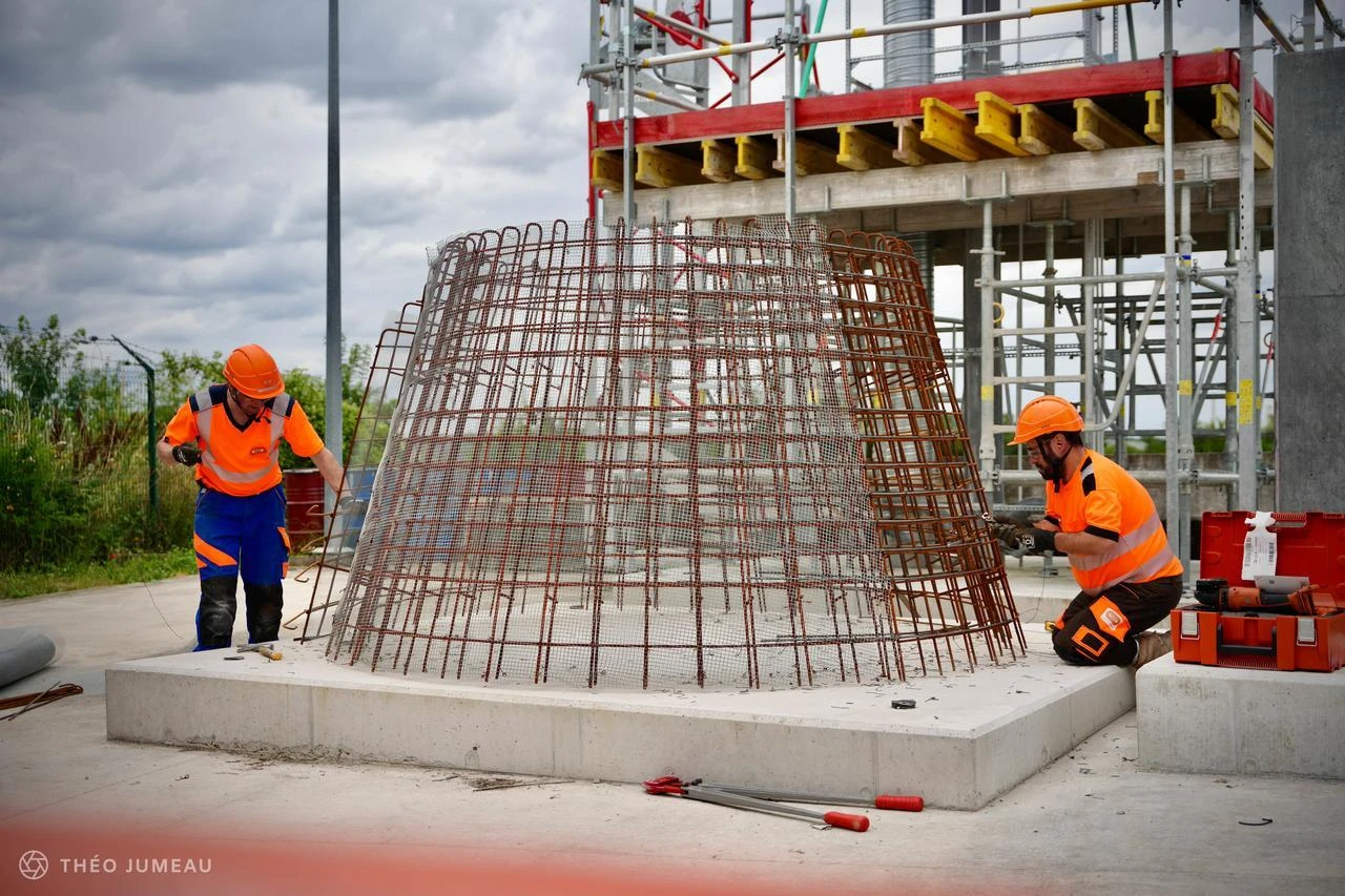 Deux ouvriers du bâtiment en tenues orange haute visibilité assemblent une grande armature métallique conique, de couleur rouille, sur une base en béton gris. Des échafaudages complexes se dressent en arrière-plan sous un ciel nuageux, indiquant un chantier de construction.