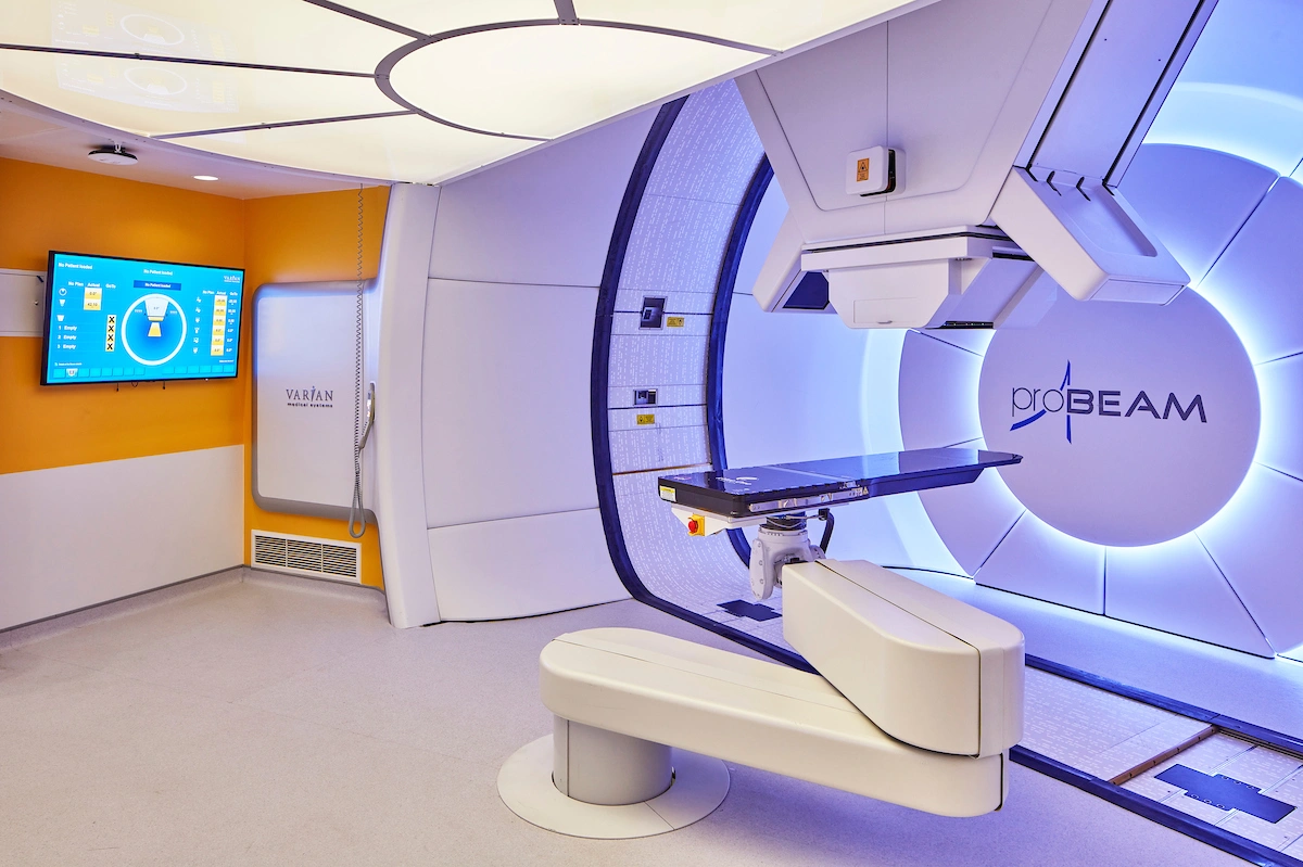 Une salle de traitement médical moderne abrite une machine de radiothérapie ProBEAM avec une table d'examen, illuminée par une lumière bleue froide. Un mur orange et blanc présente un écran affichant des données médicales et un panneau Varian Medical Systems.