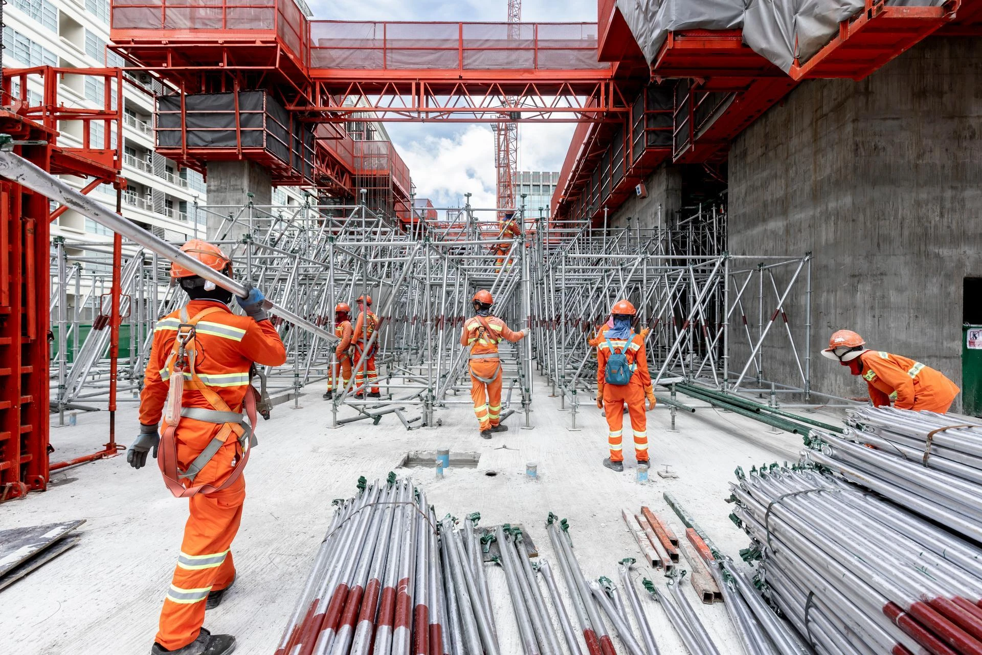 Des ouvriers de chantier en uniformes orange vif et casques de sécurité évoluent au milieu de vastes échafaudages métalliques argentés et de structures rouges, avec des piles de tuyaux au sol.
