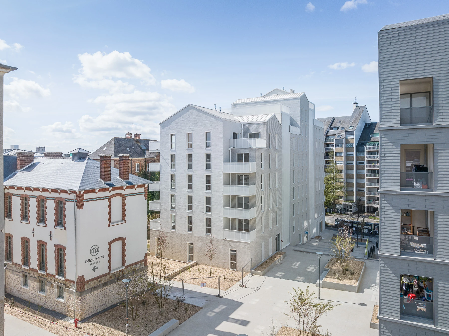 Vue aérienne d'un quartier urbain lumineux montrant des immeubles modernes blancs et gris avec balcons, jouxtant un ancien bâtiment en briques rouges et blanches, le tout sous un ciel bleu avec des aménagements paysagers au sol.