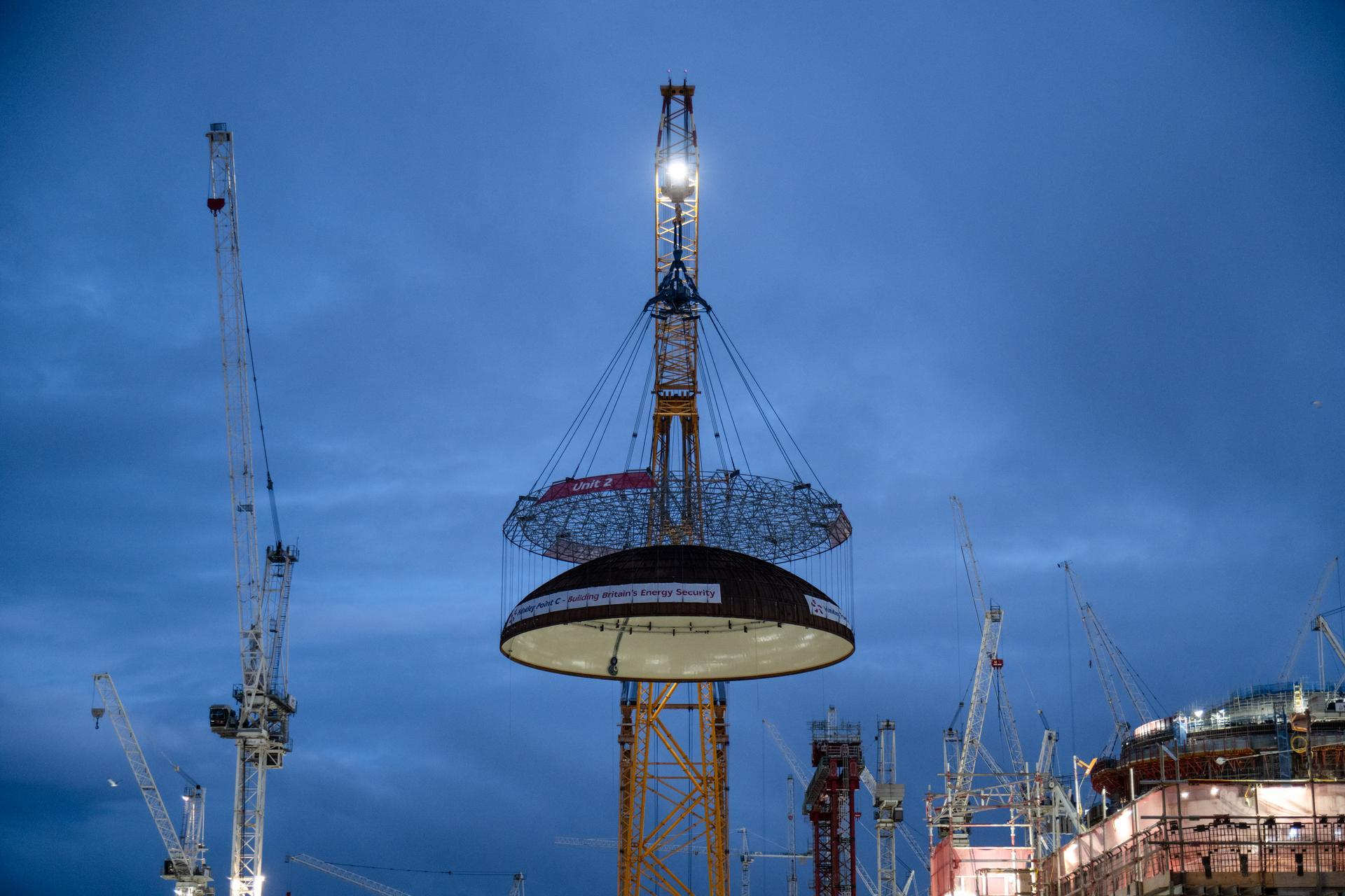 Un gigantesque grue jaune illuminée lève une grande structure en forme de dôme inversé, marquée "Unit 2" et "Hinkley Point C - Building Britain's Energy Security", sur un vaste chantier de construction sous un ciel bleu crépusculaire, entouré d'autres grues.