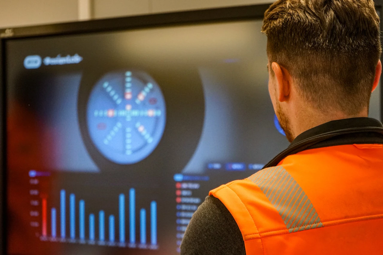 Vue de dos d'un homme portant un gilet orange haute visibilité, observant un grand écran affichant des visualisations de données techniques, dont un diagramme circulaire bleu et des graphiques à barres bleus et rouges.