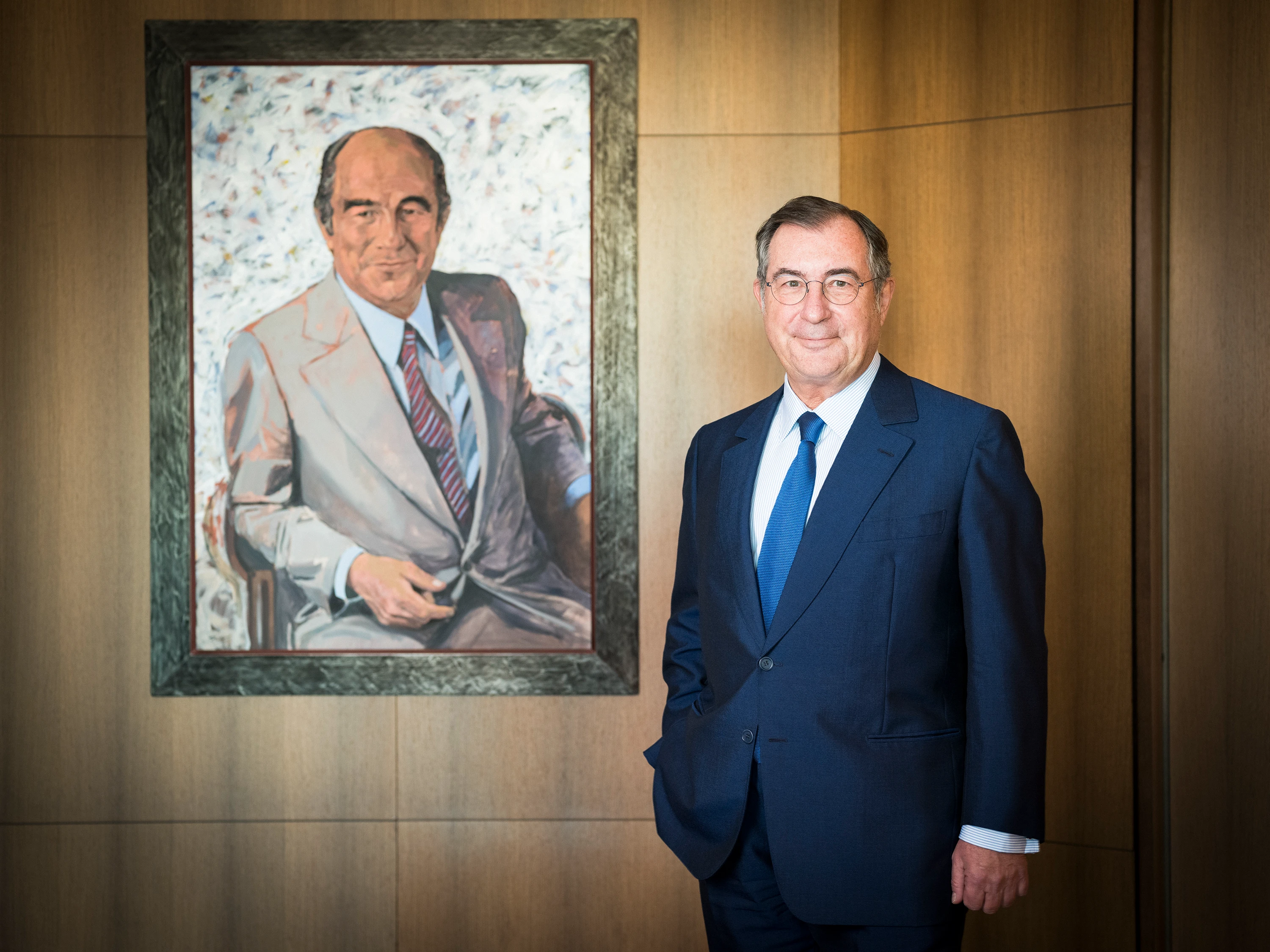 Un homme souriant, vêtu d'un costume bleu foncé et portant des lunettes, se tient à côté d'un portrait encadré représentant un homme en costume clair sur fond abstrait, le tout devant un mur en bois chaleureux.