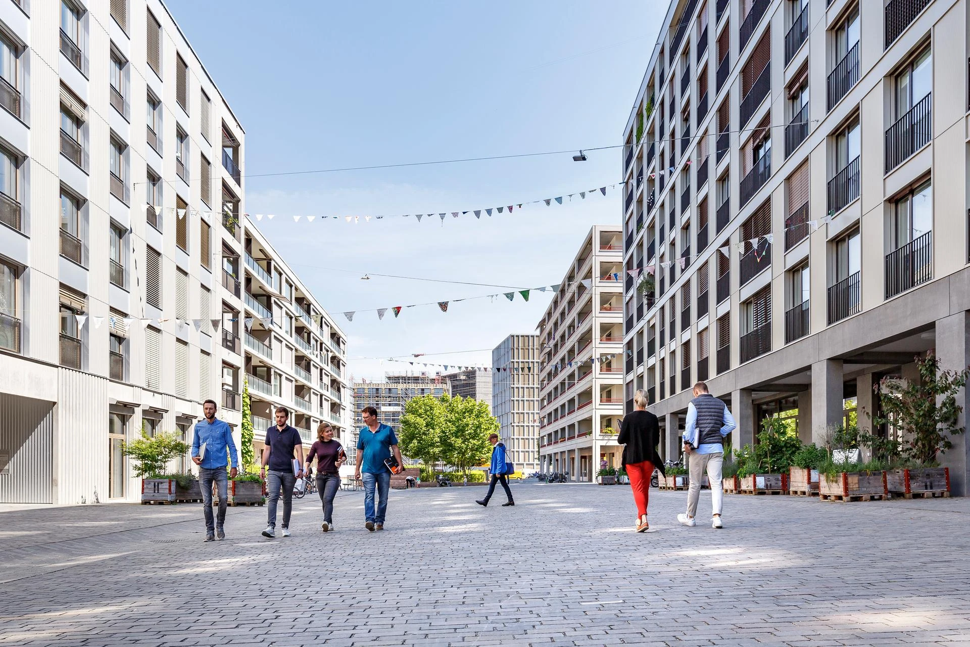 Plusieurs personnes traversent une place urbaine pavée bordée d'immeubles modernes clairs et de verdure, sous un ciel bleu orné de guirlandes, avec des constructions en arrière-plan.