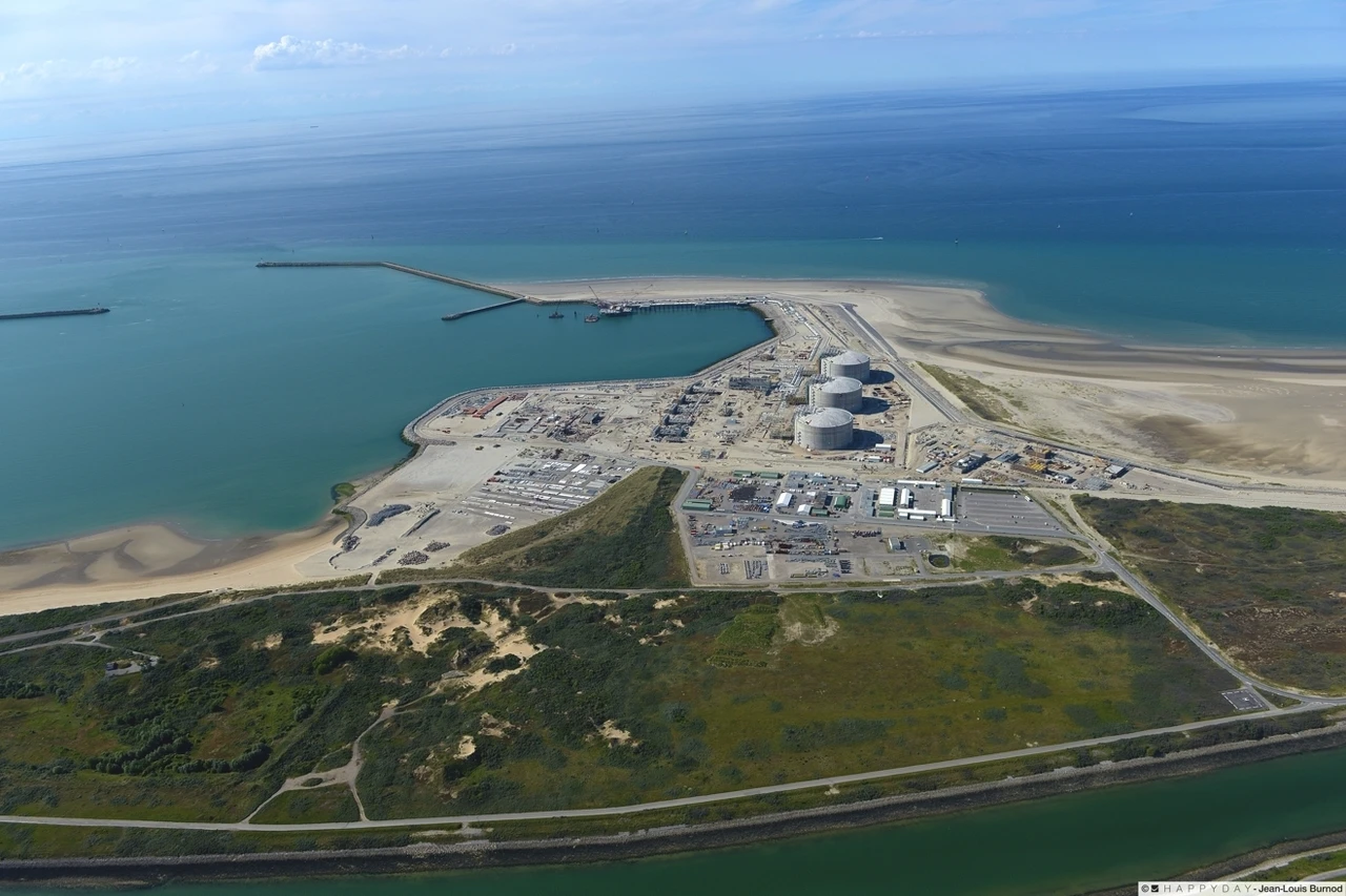 Vue aérienne d'un complexe industriel côtier avec de grands réservoirs cylindriques blancs et un port aux eaux turquoise, s'étendant sur une langue de terre sablonneuse. Il est bordé par l'océan bleu profond, des plages, des dunes vertes et un canal sinueux.
