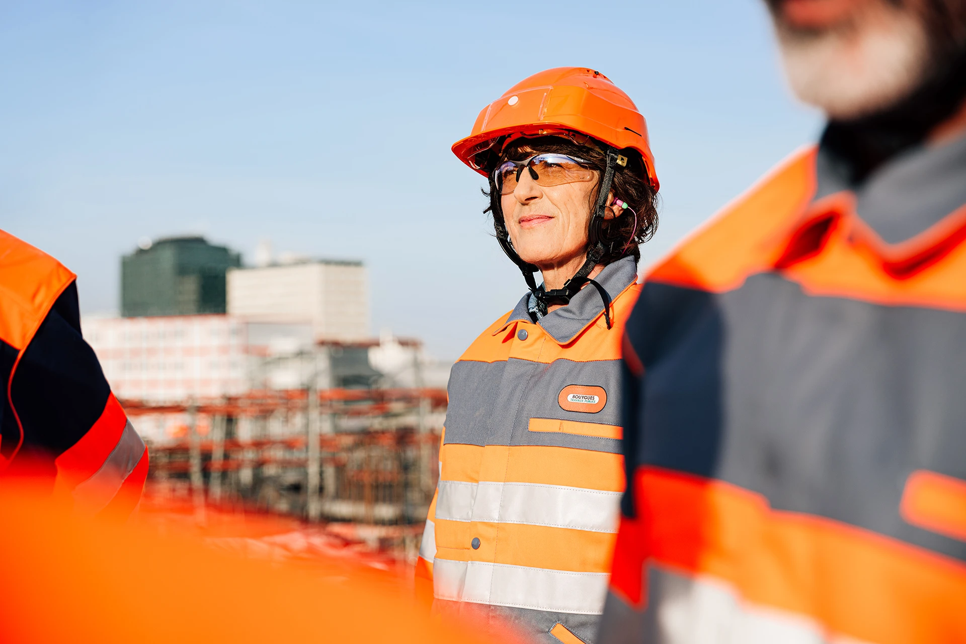 Femme souriante en casque de sécurité orange, lunettes de protection et veste de travail grise et orange à bandes réfléchissantes avec le logo Bouygues Construction, sur un chantier sous un ciel bleu clair.