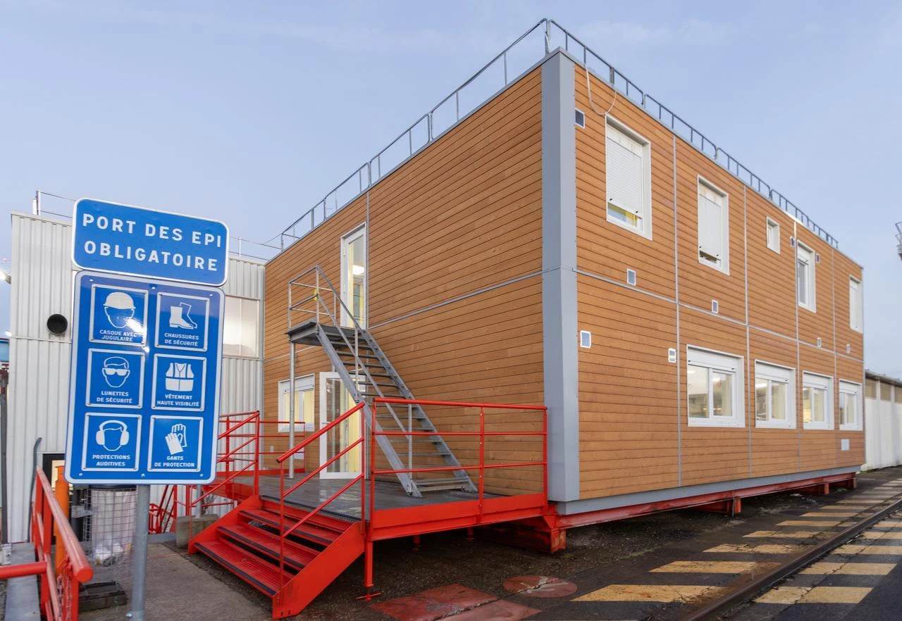 Un bâtiment modulaire moderne de deux étages avec une façade marron clair et un escalier métallique rouge menant à l'entrée. Un panneau de sécurité bleu bien visible, « PORT DES EPI OBLIGATOIRE », avec des pictogrammes indiquant les EPI obligatoires, se trouve au premier plan sur un site industriel.
