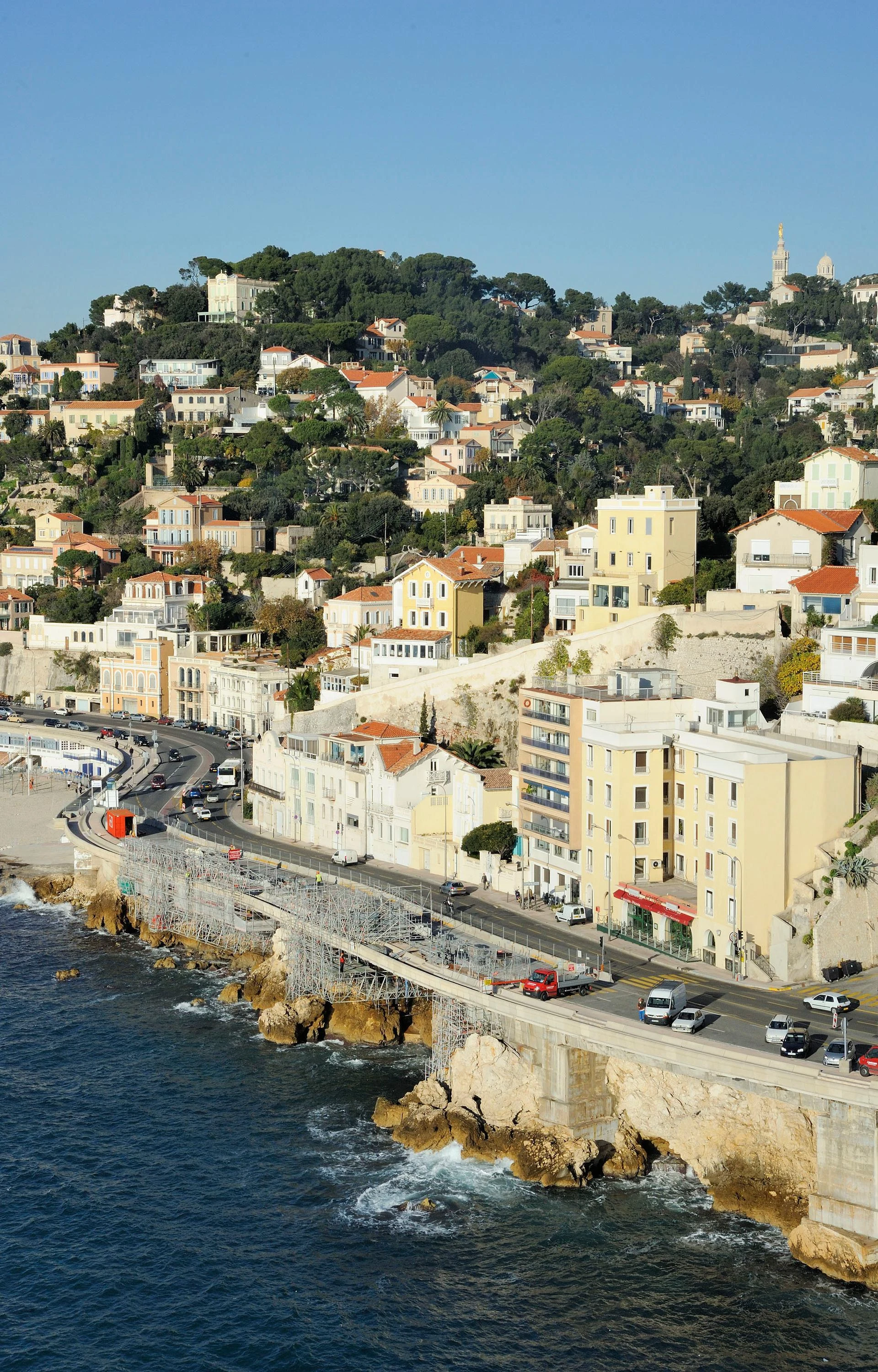 Vue aérienne d'une ville côtière méditerranéenne ensoleillée, avec une route sinueuse longeant la mer bleue et des bâtiments clairs aux toits rouges à flanc de colline. Des échafaudages sont visibles le long du rivage rocheux, et la basilique Notre-Dame de la Garde domine l'horizon au sommet de la colline.