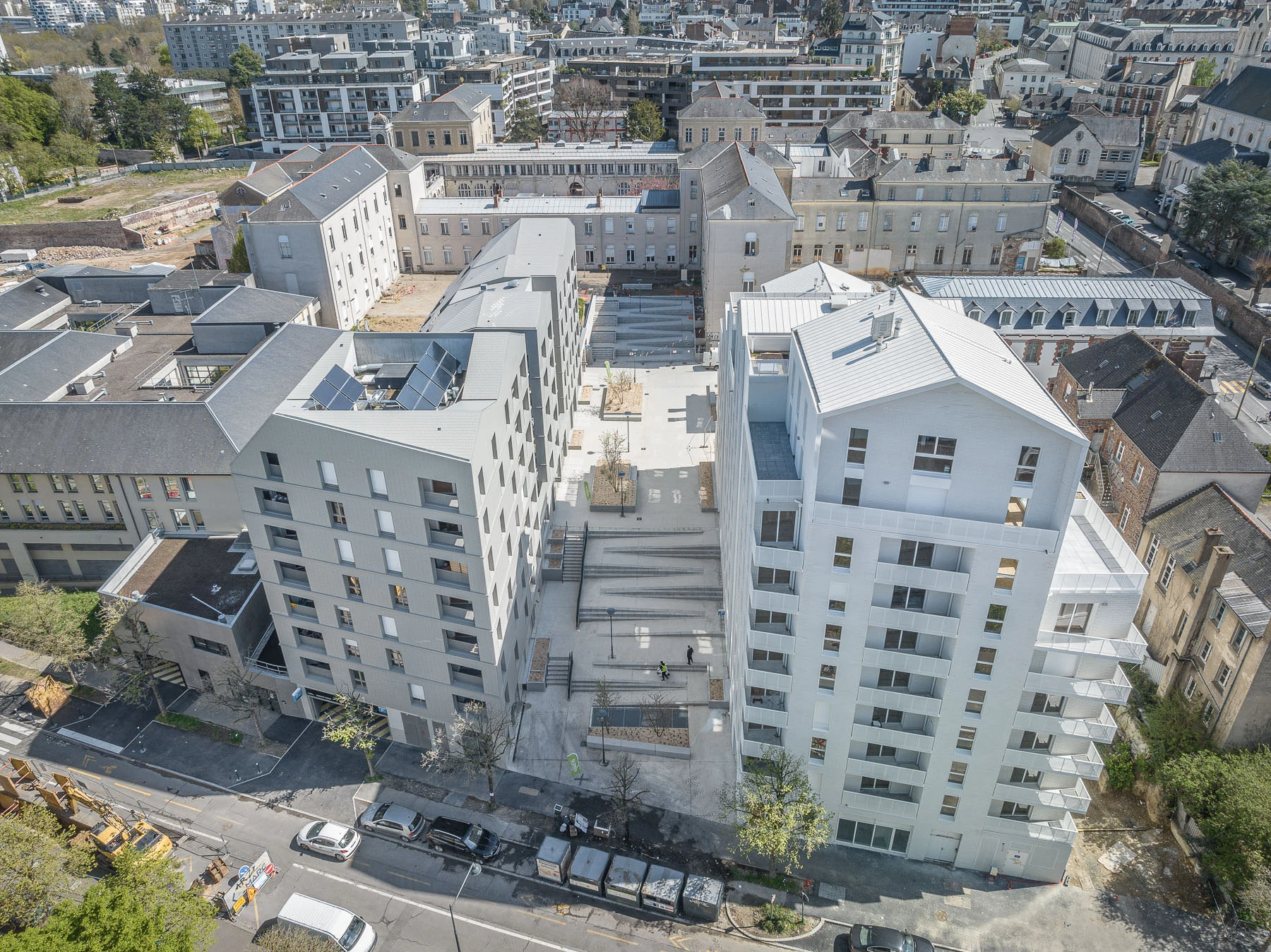 Une vue aérienne montre un développement urbain moderne comprenant deux immeubles résidentiels angulaires gris clair, dont l'un est équipé de panneaux solaires, surplombant une place en béton en terrasse. Ils sont nichés dans un paysage urbain dense composé de bâtiments anciens, de rues et d'espaces verts dispersés.