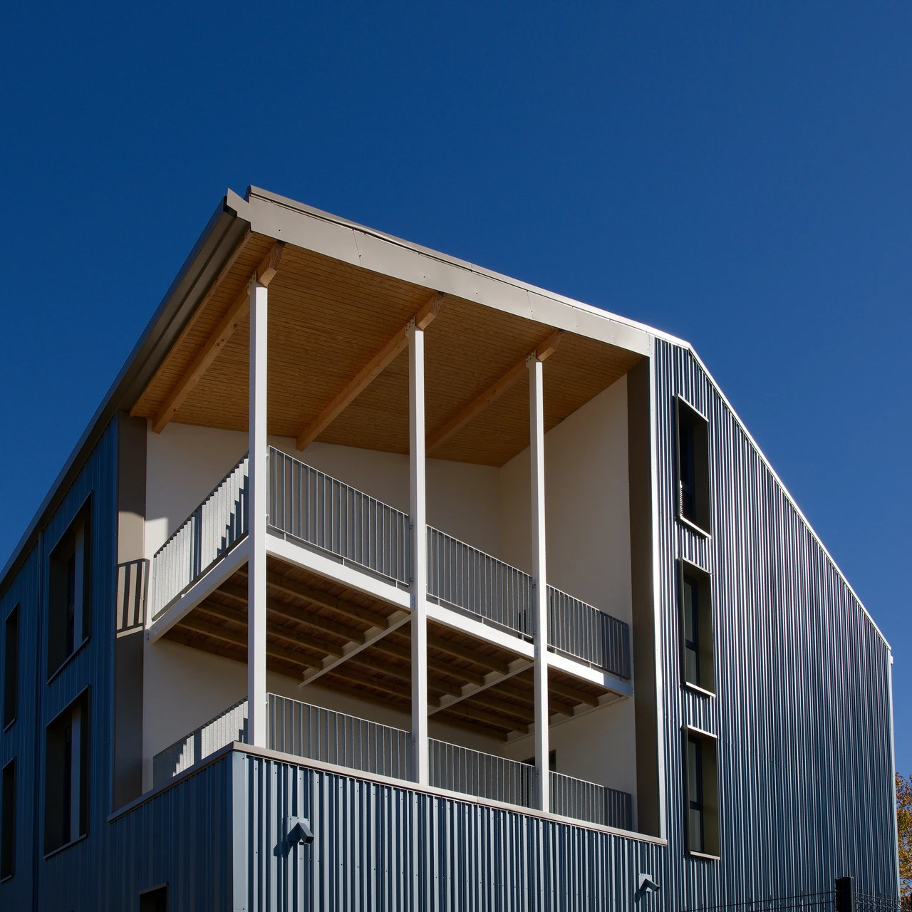 Façade d'un bâtiment moderne avec des balcons superposés, des supports blancs, des rambardes métalliques et un revêtement en panneaux métalliques verticaux sous un ciel bleu.