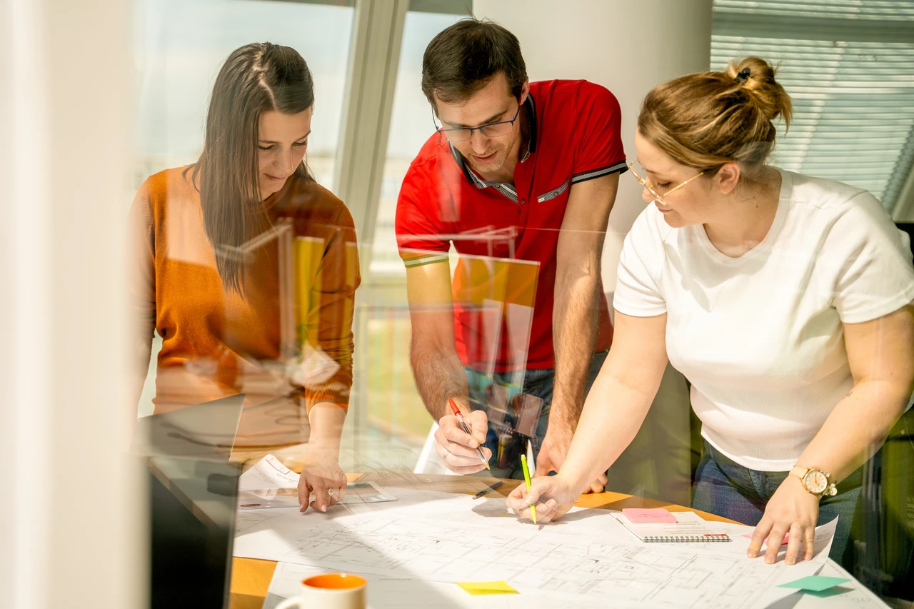 Trois professionnels collaborent autour d'une table, examinant des plans et prenant des notes avec des stylos dans un bureau lumineux.