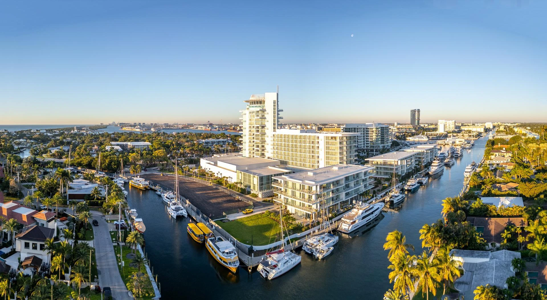 Aerial panorama of a vibrant coastal city showcasing numerous yachts and boats docked along a network of canals. Modern white buildings, lush palm trees, and residential homes line the waterways under a clear blue sky.