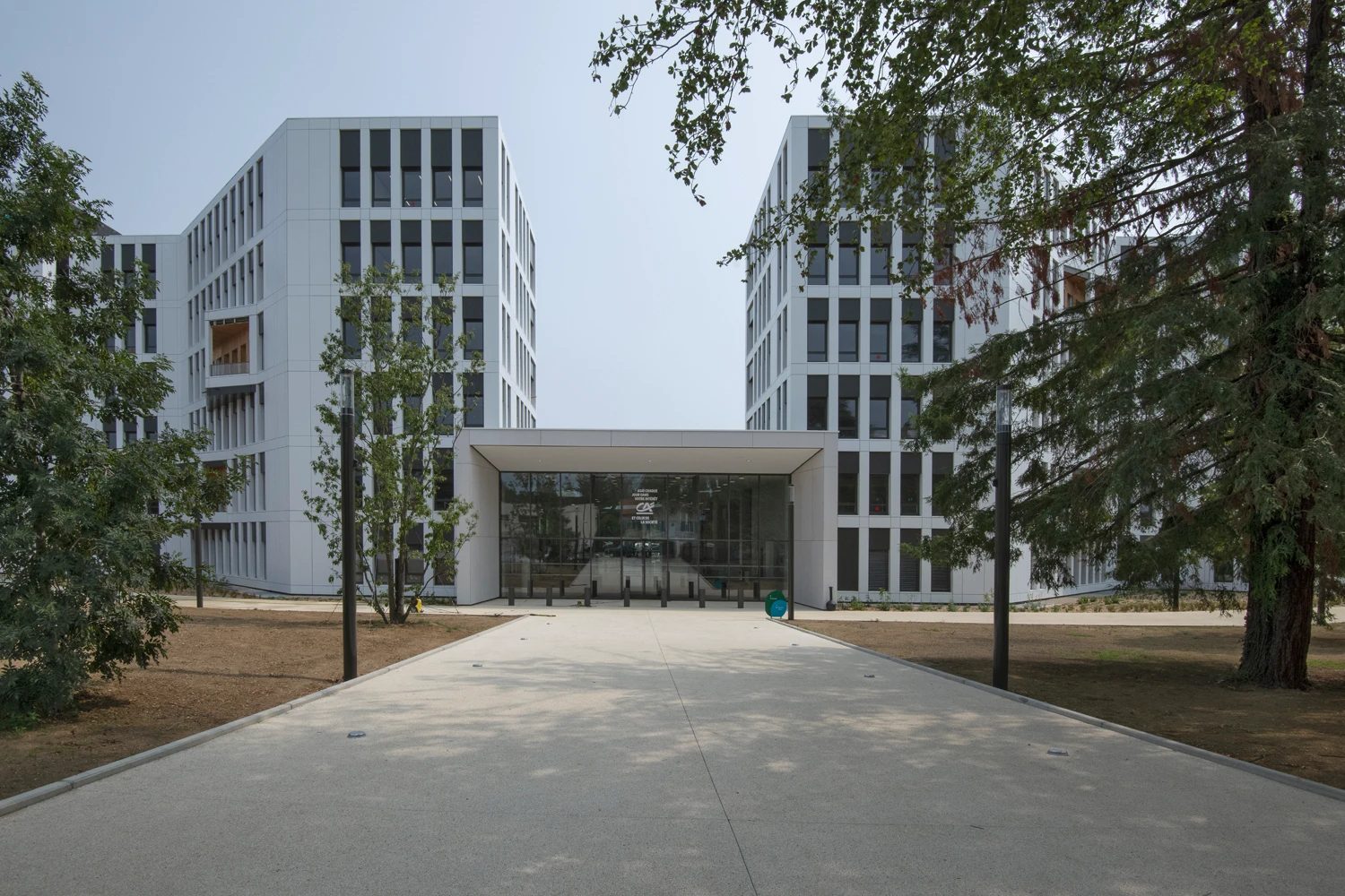 Deux immeubles modernes blancs et symétriques avec des fenêtres sombres sont reliés par une entrée vitrée centrale affichant le logo Crédit Agricole, bordés d'arbres et d'une allée pavée.