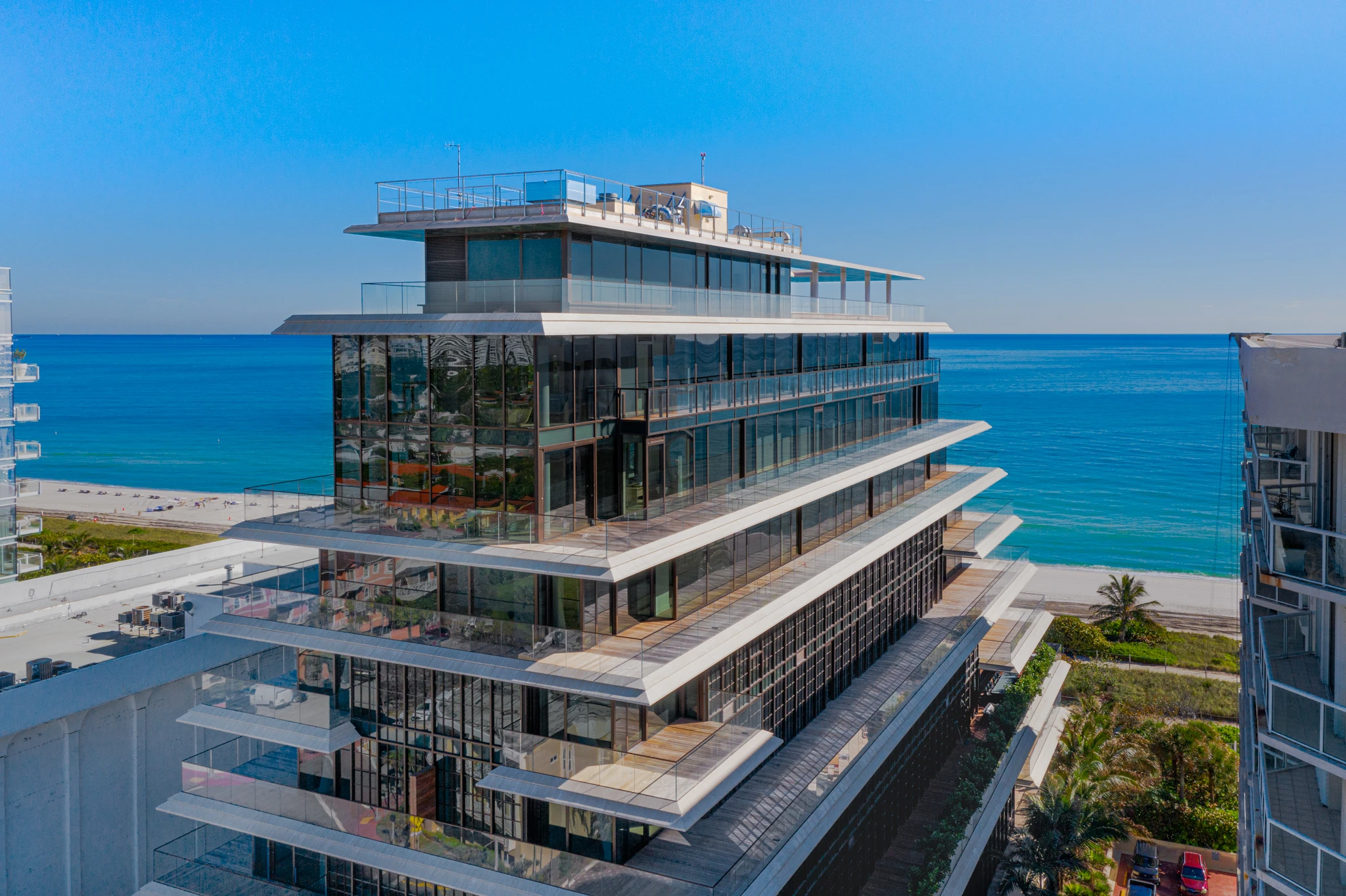 Immeuble moderne de grande hauteur avec une façade en verre réfléchissant et des balcons blancs en gradins donnant sur un océan bleu vif, une plage de sable blanc et un ciel clair.