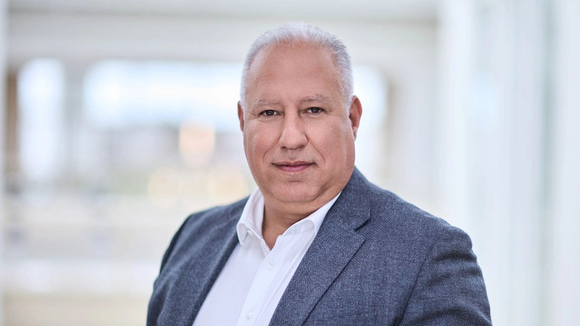 Portrait d'un homme souriant aux cheveux gris, vêtu d'une chemise blanche et d'une veste texturée bleu-gris, devant un arrière-plan intérieur lumineux et flou.