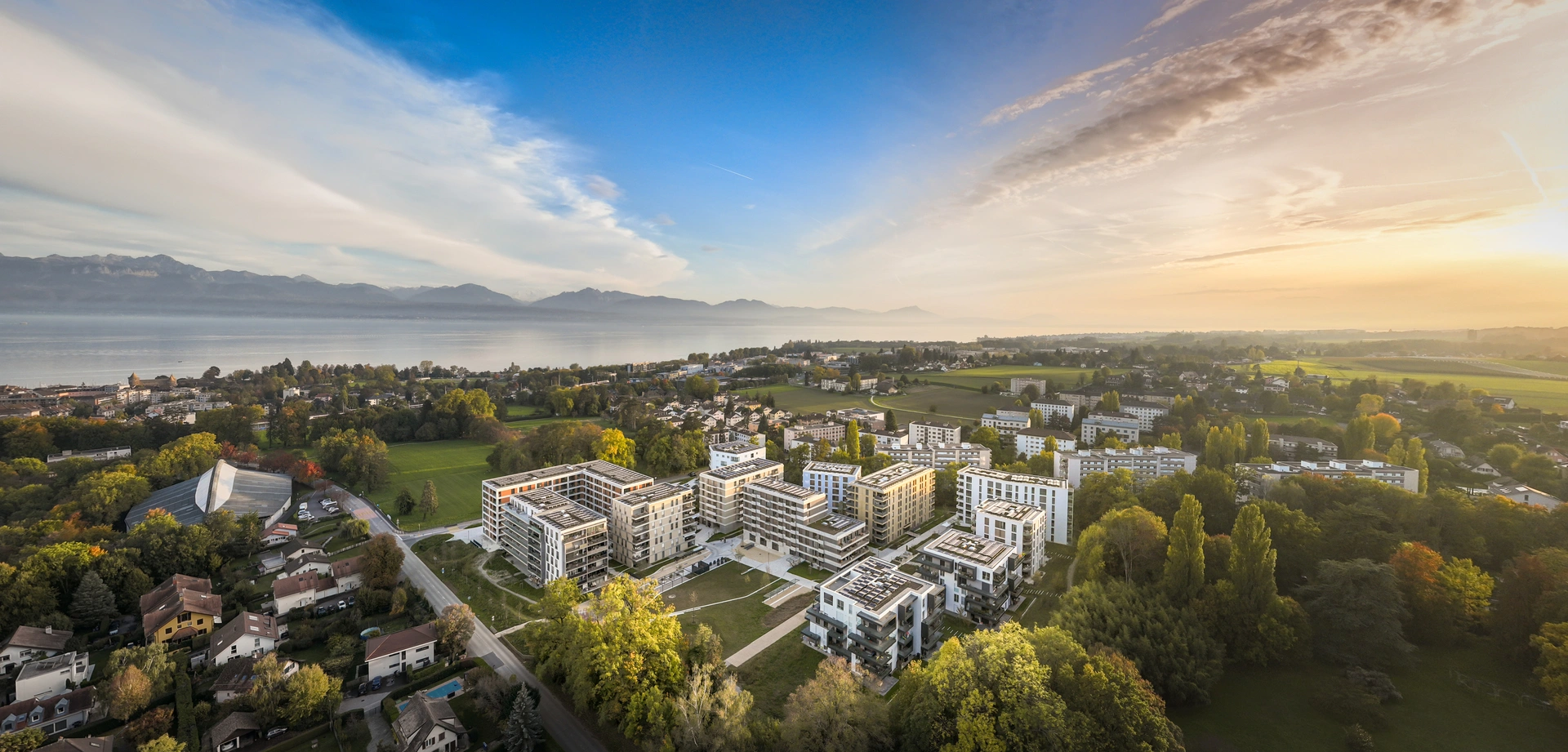 Vue aérienne d'un quartier moderne avec des immeubles clairs et des toits gris, entouré de verdure. Le panorama s'étend sur un grand lac, une ville et des montagnes à l'horizon, sous un ciel bleu clair baigné d'une lumière chaude de lever ou coucher de soleil.