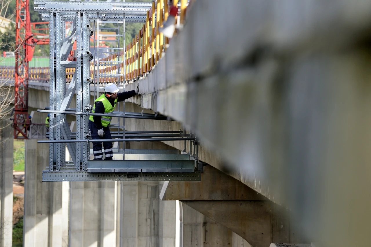 Un ouvrier du bâtiment inspecte le dessous d'un pont en béton depuis une plateforme de travail temporaire.