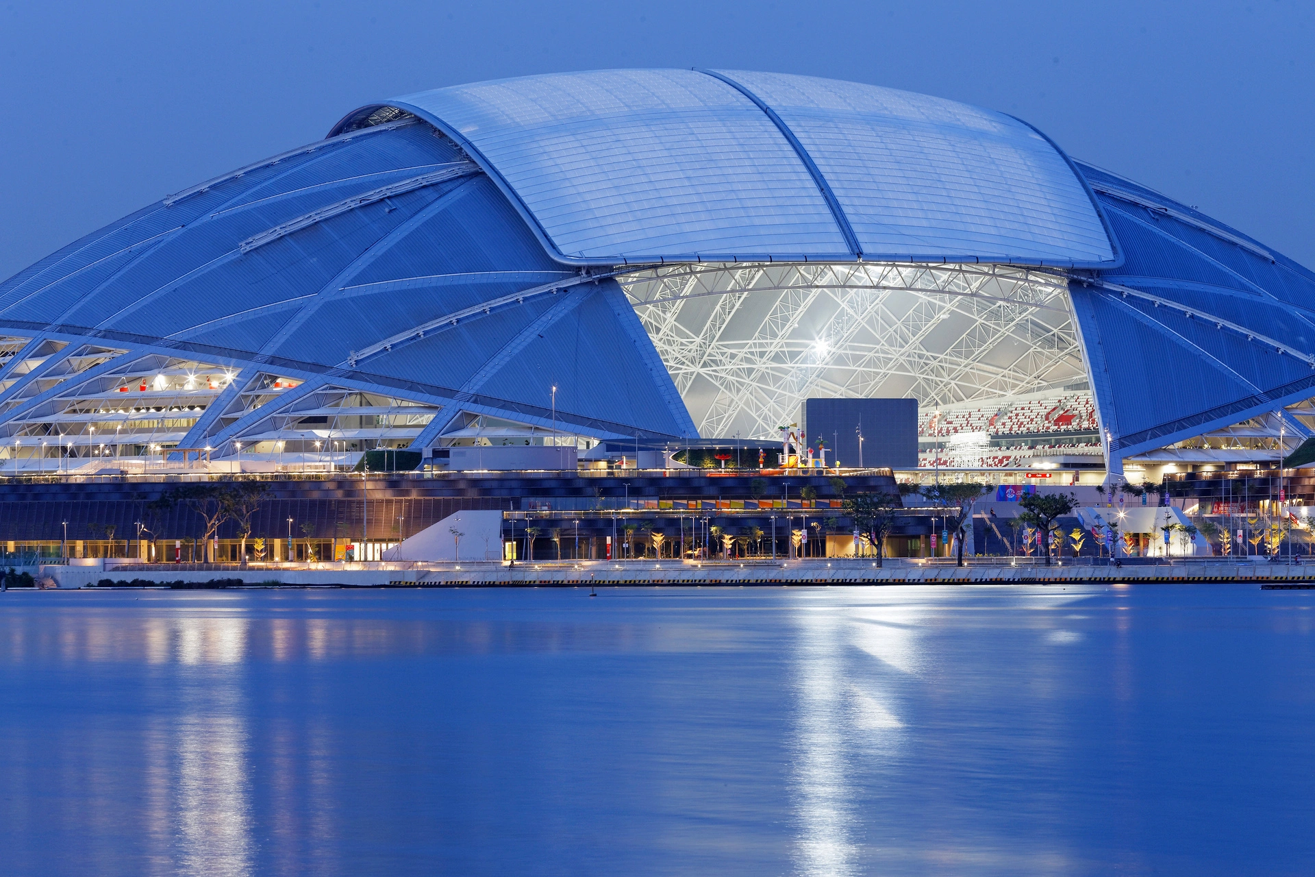 Un imposant stade moderne, reconnaissable à son toit dôme partiellement ouvert et sa structure complexe, brille de lumières vives blanches et jaunes au crépuscule, se reflétant sur l'eau bleue. L'intérieur révèle des sièges rouges et blancs sous la charpente lumineuse.