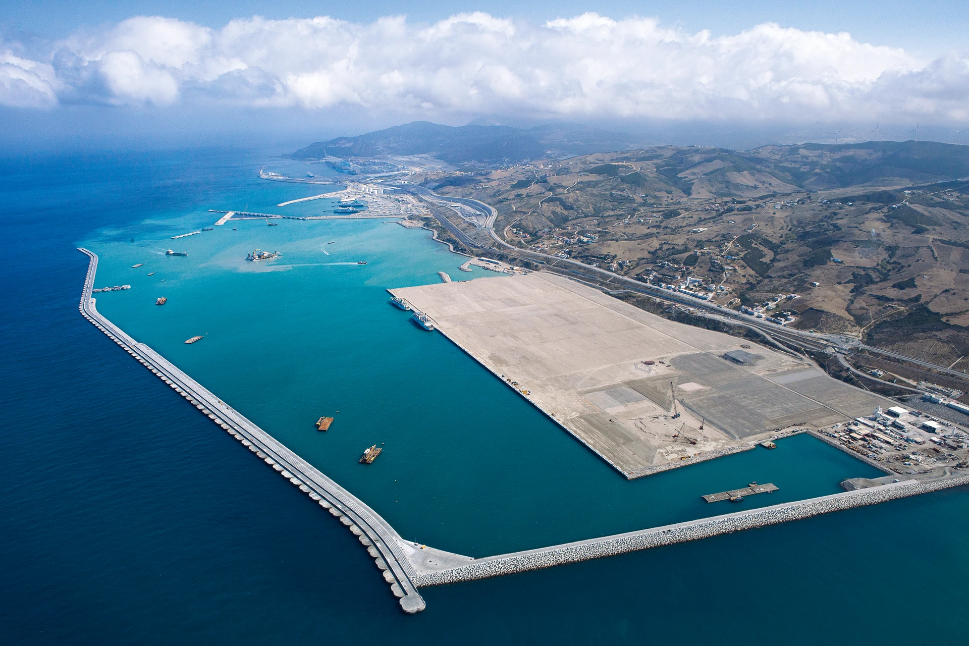 Vue aérienne d'un grand port moderne avec un long brise-lames incurvé séparant les eaux turquoise du port de la mer d'un bleu profond. De vastes zones portuaires claires sont entourées de collines arides, de routes côtières et de nuages blancs.