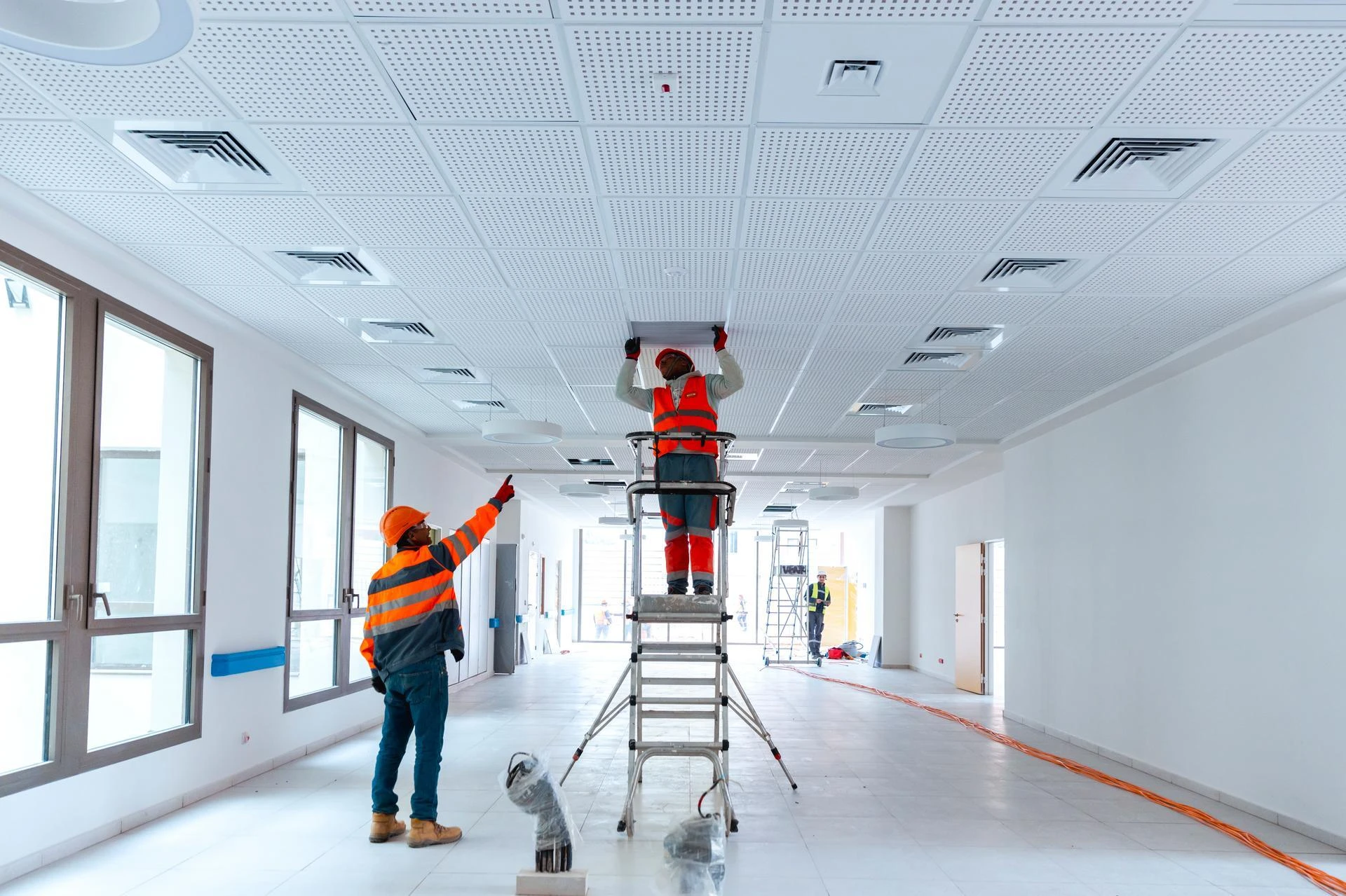 Deux ouvriers du bâtiment, vêtus d'équipements de sécurité orange, installent des panneaux au plafond perforé blanc d'un couloir lumineux, l'un sur une échelle et l'autre pointant vers le haut.