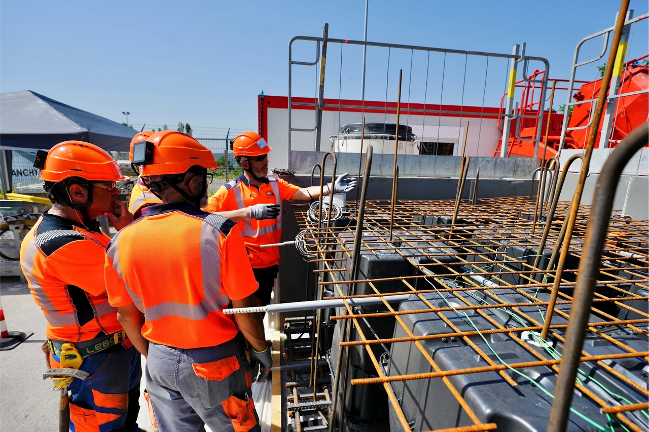 Quatre ouvriers en orange vif et casques de sécurité inspectent une structure en acier armé et coffrages modulaires noirs sur un chantier ensoleillé. Un ouvrier au centre pointe et explique les détails de la construction, sous un ciel bleu clair.