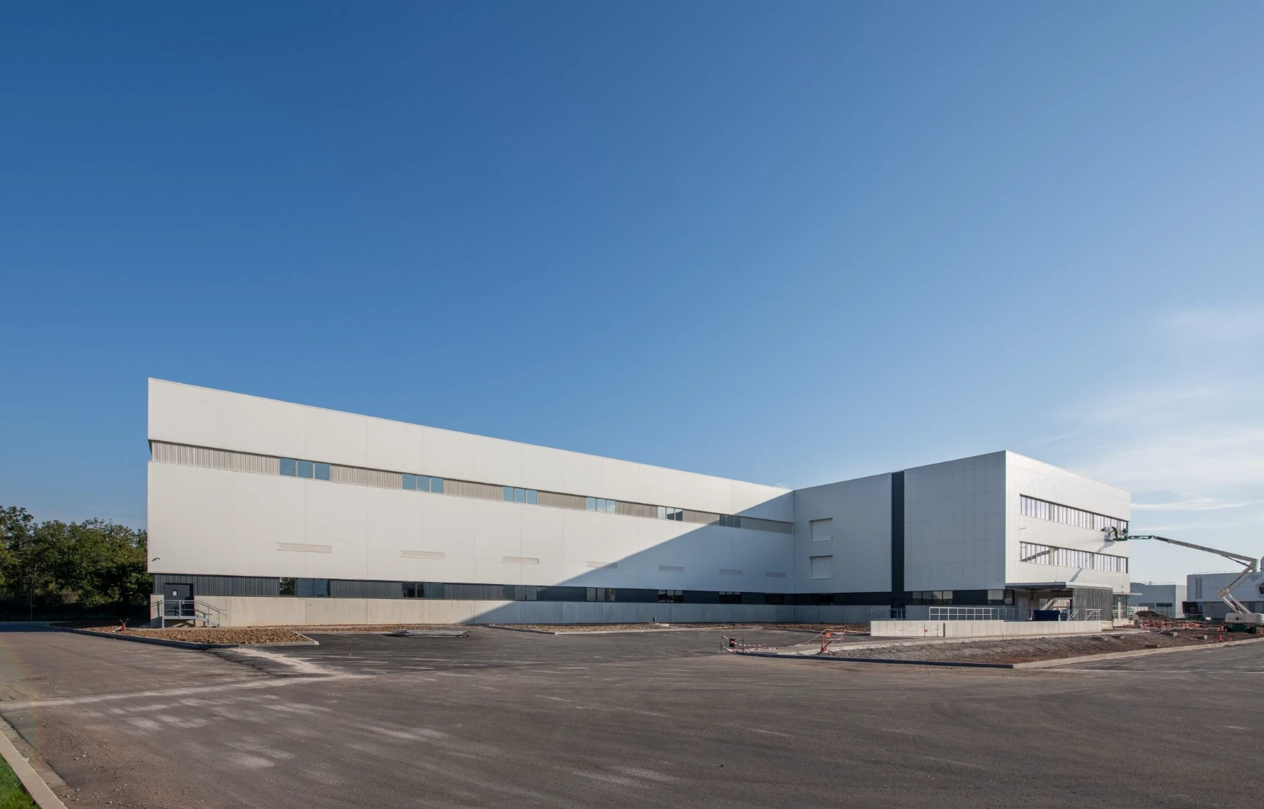 Grand bâtiment industriel ou commercial moderne aux façades blanches et grises, avec des lignes épurées et de nombreuses fenêtres, sous un ciel bleu. Un vaste parking asphalté et une nacelle élévatrice près de l'entrée suggèrent un site récent ou en cours d'aménagement.