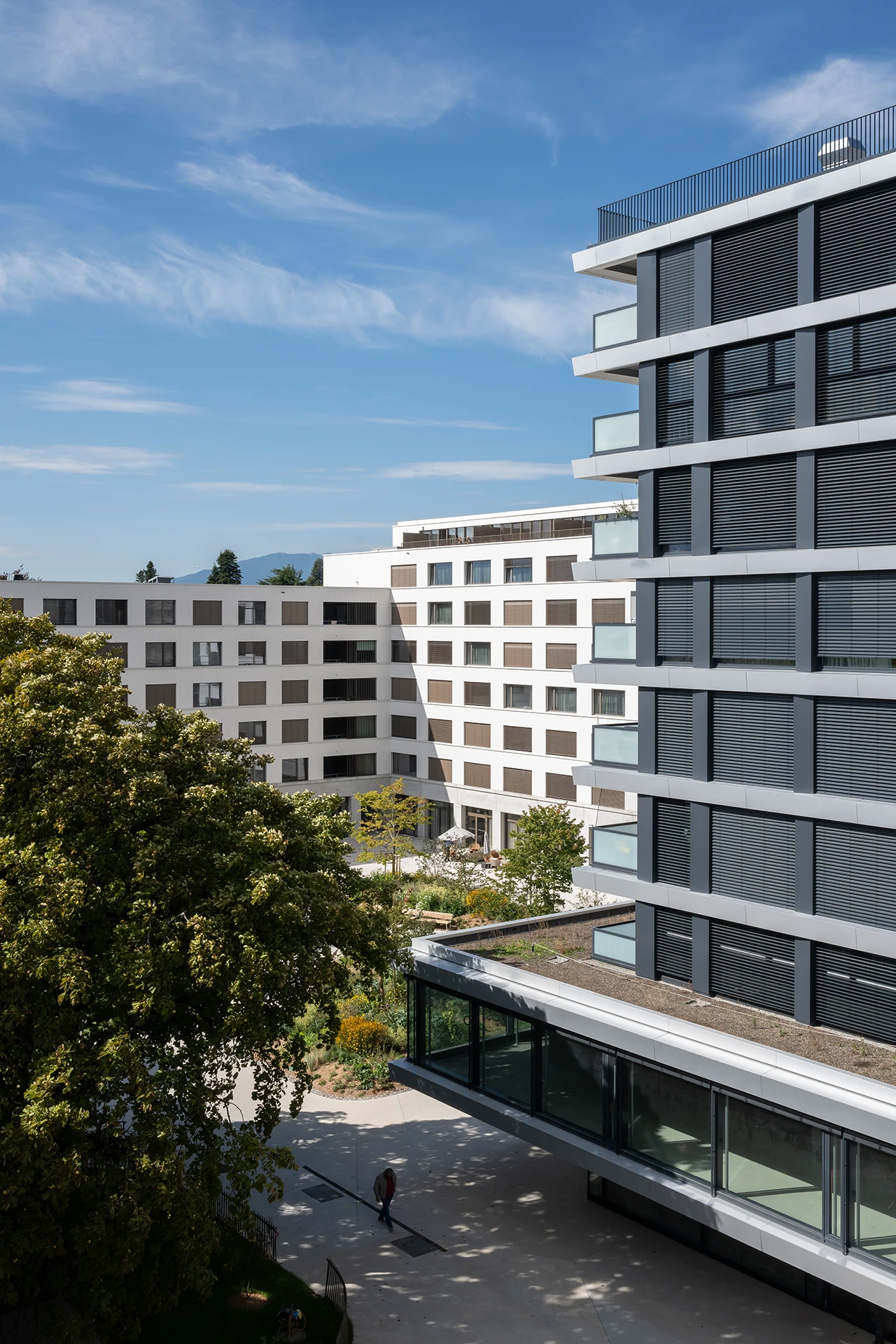Complexe architectural moderne avec un bâtiment principal gris foncé à stores sur la droite et des bâtiments blancs en arrière-plan encadrant un jardin luxuriant avec des arbres, des parterres de fleurs et une personne marchant, sous un ciel bleu parsemé de nuages.