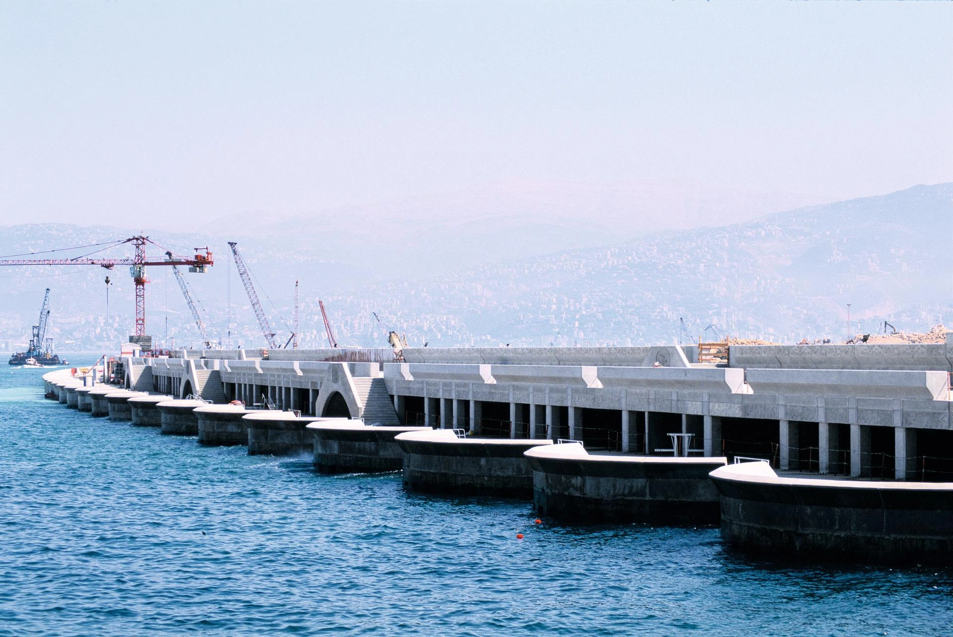 Un long brise-lames ou quai en béton clair en construction s'étend dans des eaux bleues profondes, avec des grues rouges et grises visibles derrière lui. Des montagnes brumeuses et un paysage urbain apparaissent à l'horizon sous un ciel pâle.