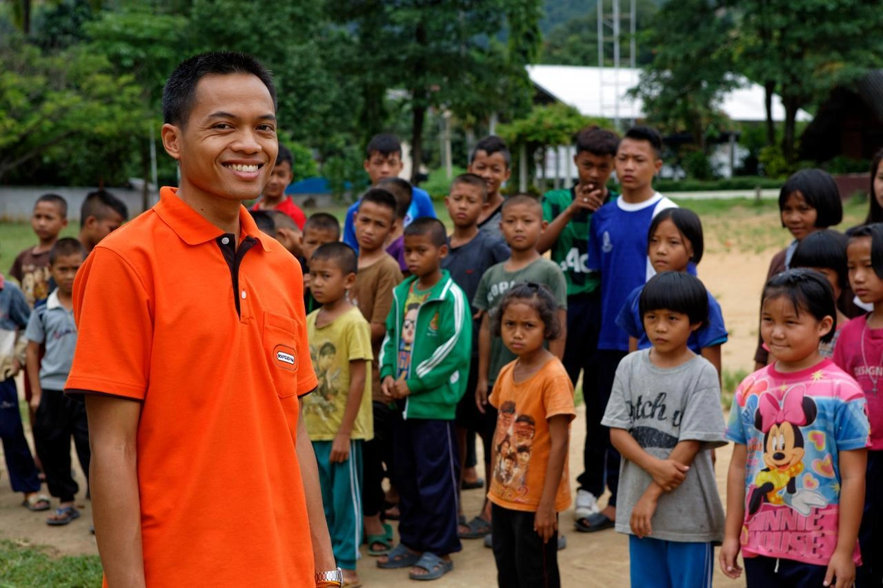 Un homme asiatique souriant, vêtu d'un polo orange vif, se tient au premier plan devant un groupe flou d'enfants aux vêtements colorés dans un cadre extérieur verdoyant.