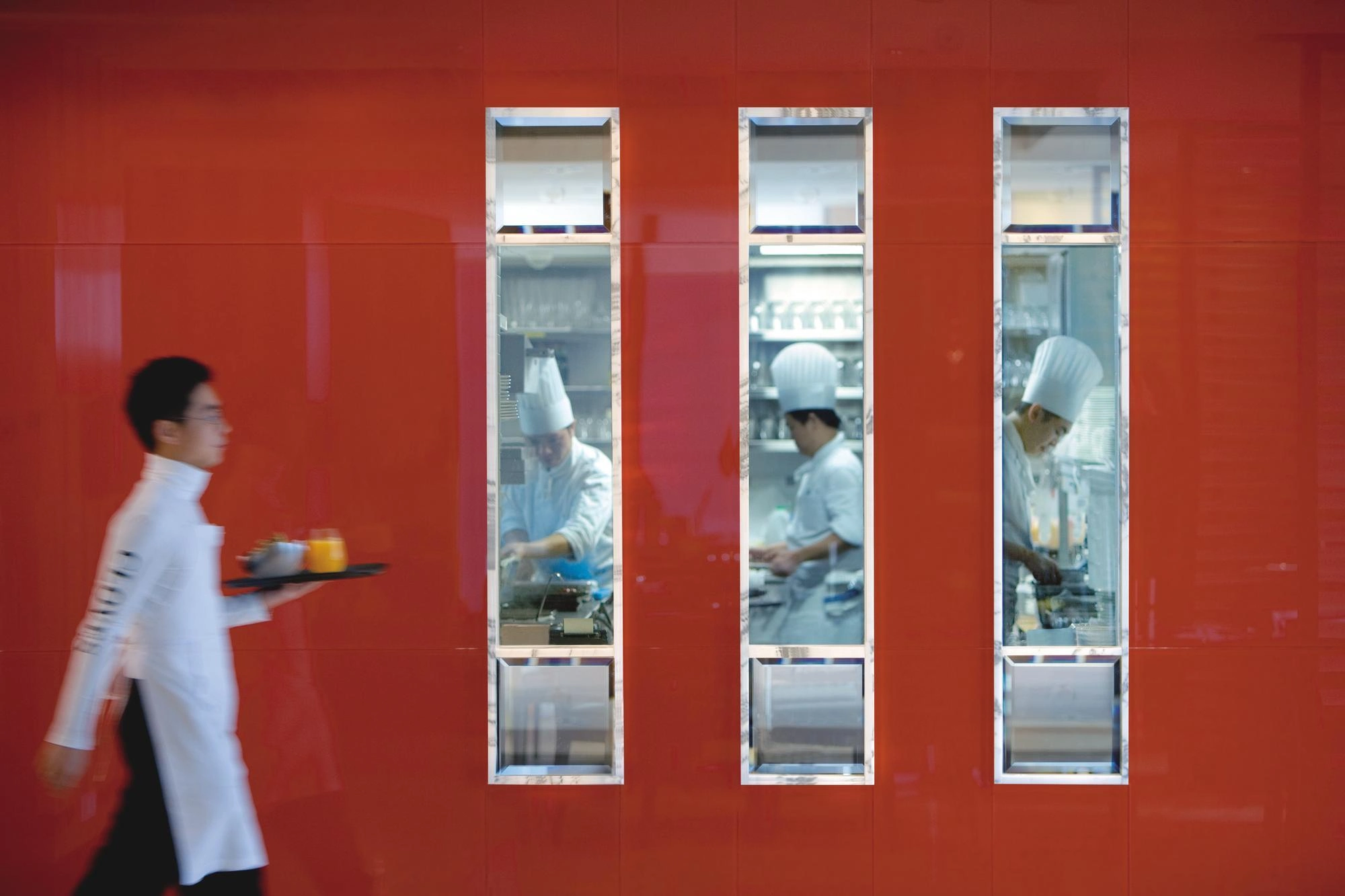 Un serveur portant un plateau passe devant trois fenêtres montrant des chefs dans une cuisine sur un mur rouge.