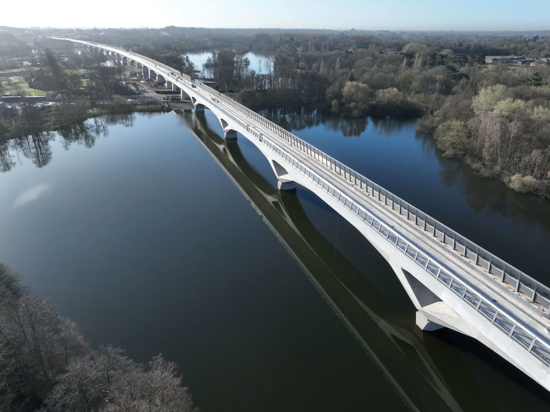 Vue aérienne d'un long pont ferroviaire blanc et moderne avec des arches, traversant une rivière sombre et reflétant dans l'eau, entouré de berges boisées sous un ciel clair et brumeux.