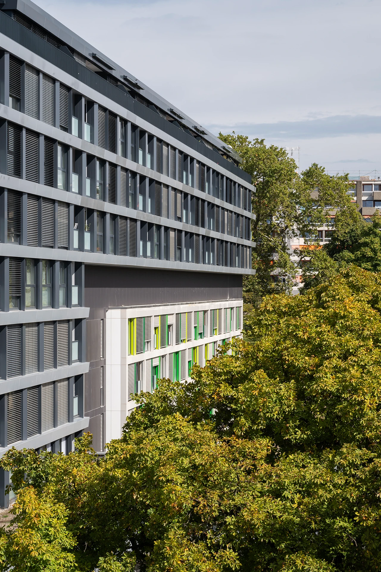 Un bâtiment moderne gris foncé avec des persiennes horizontales et un bâtiment blanc aux fenêtres bordées de vert vif, encadrés par des feuillages denses et verdoyants sous un ciel nuageux.