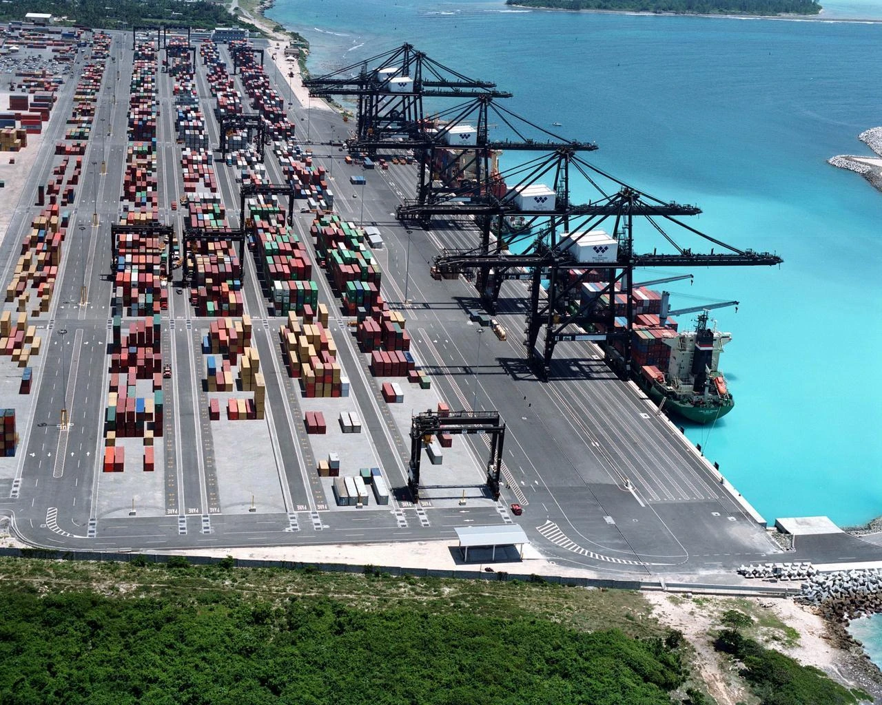 Vue aérienne d'un grand port à conteneurs où des milliers de conteneurs colorés sont empilés le long de quais gris, desservis par de massives grues noires chargeant un cargo vert dans des eaux turquoise.