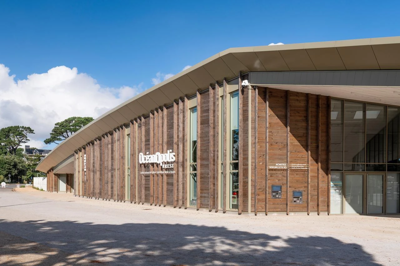 Vue extérieure du bâtiment Océanopolis Brest sous un ciel bleu, caractérisé par son architecture moderne avec des lattes de bois verticales et de grandes fenêtres.