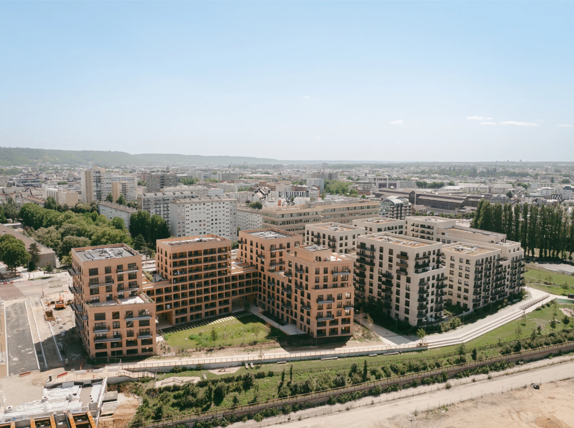 Vue aérienne de plusieurs immeubles résidentiels modernes beige et terracotta avec balcons et toits plats, entourés d'espaces verts. Au premier plan, un chantier de construction côtoie une ville étendue sous un ciel bleu dégagé.