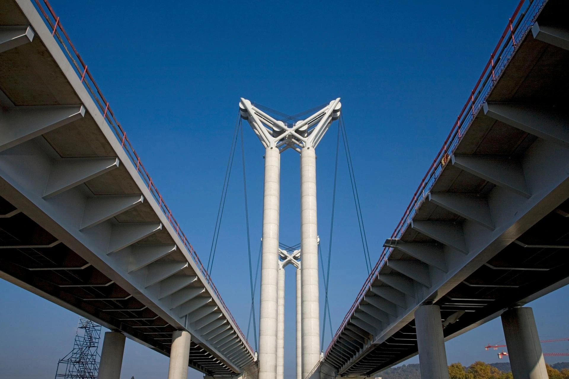 Vue en contre-plongée d'un pont à haubans moderne en béton gris, avec des balustrades rouges, dont les tabliers convergent vers deux pylônes centraux massifs supportant des structures blanches en X et V sous un ciel bleu éclatant.