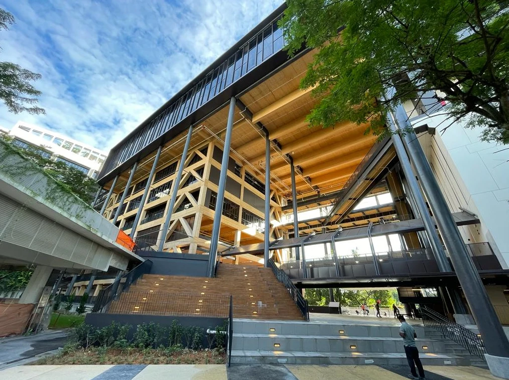 Un bâtiment moderne et imposant affiche une structure externe en bois clair visible et des panneaux sombres, avec de grandes baies vitrées, le tout sous un ciel bleu nuageux. Un large escalier en bois mène à son entrée principale, complété par des passerelles et de la verdure environnante.